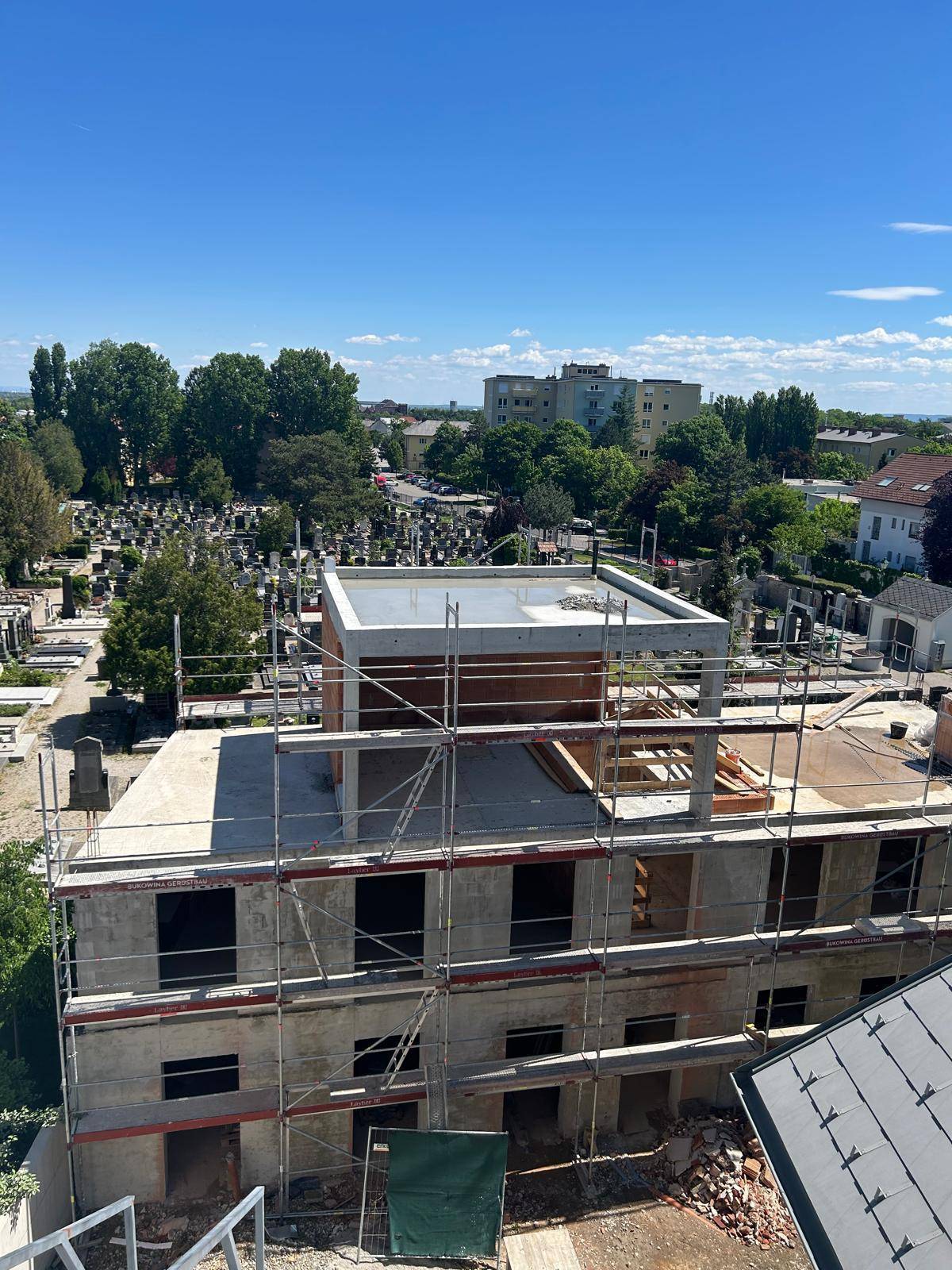Neubau im Rohbau mit Gerüst und Blick auf die umliegende Stadtlandschaft und einen Friedhof.