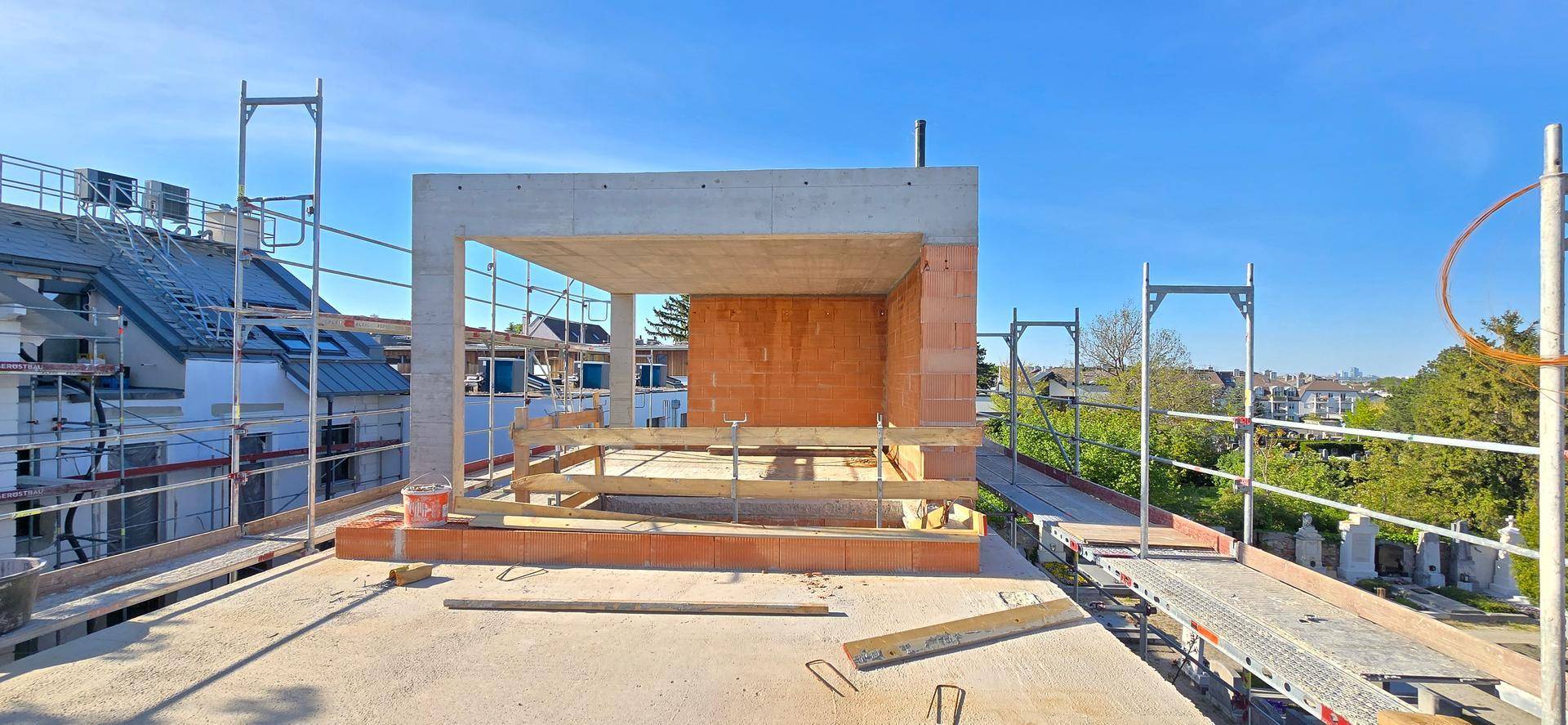 Baustelle einer Dachterrasse mit Gerüst und Blick auf umliegende Gebäude und Grünflächen.