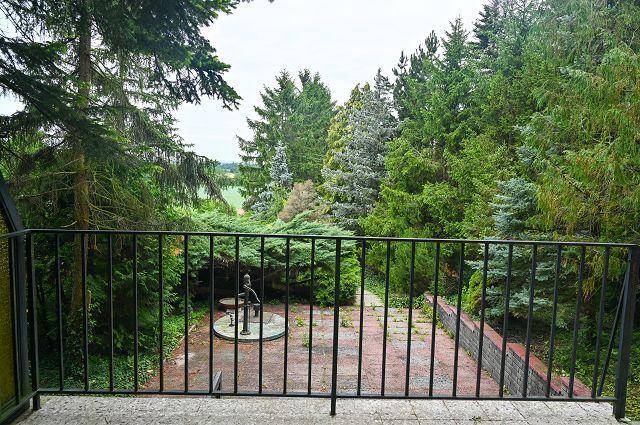 Blick vom Balkon auf eine gepflasterte Fläche mit Brunnen, umgeben von üppigem Grün und Bäumen.