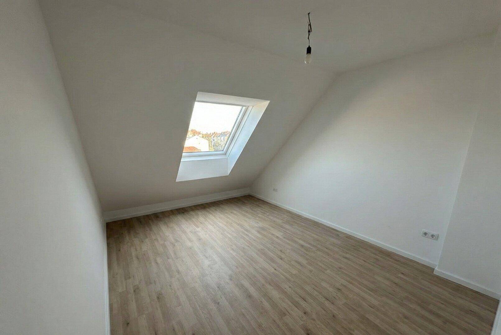 Geräumiger Dachgeschossraum mit schrägen Wänden, einem Dachfenster und hellem Laminatboden.
