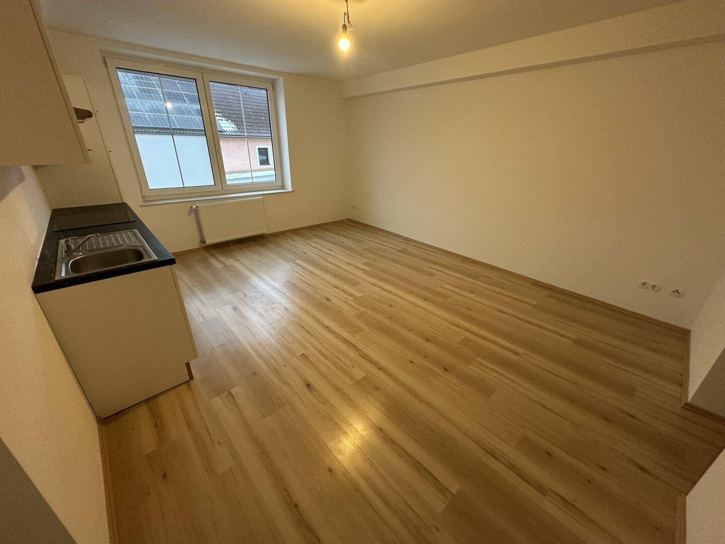 Geräumiger Wohnbereich mit hellem Holzfußboden und großem Fenster, das viel Tageslicht hereinlässt.