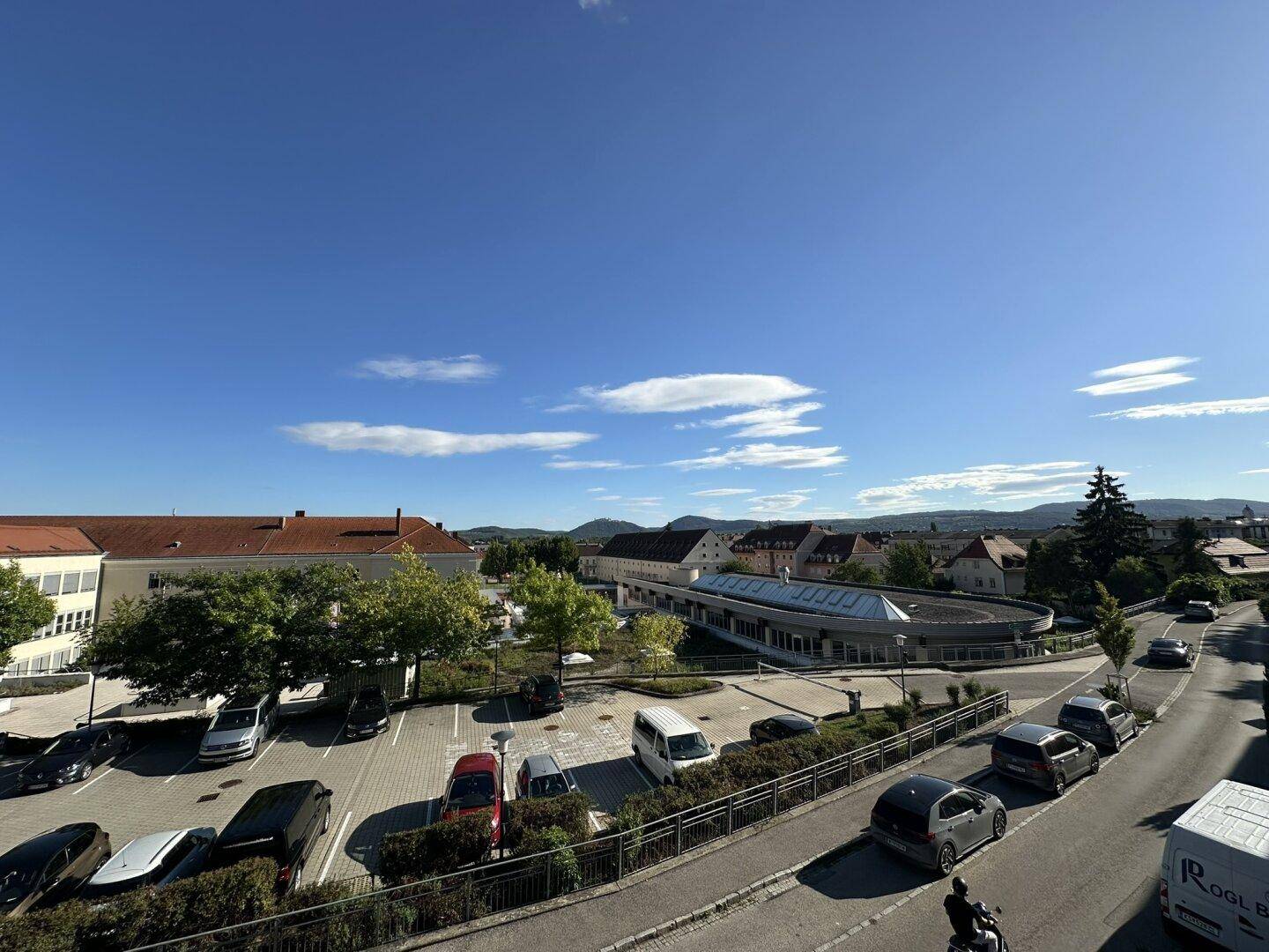 Panoramablick über die Stadt mit Gebäuden, Parkplätzen und hügeliger Landschaft im Hintergrund.