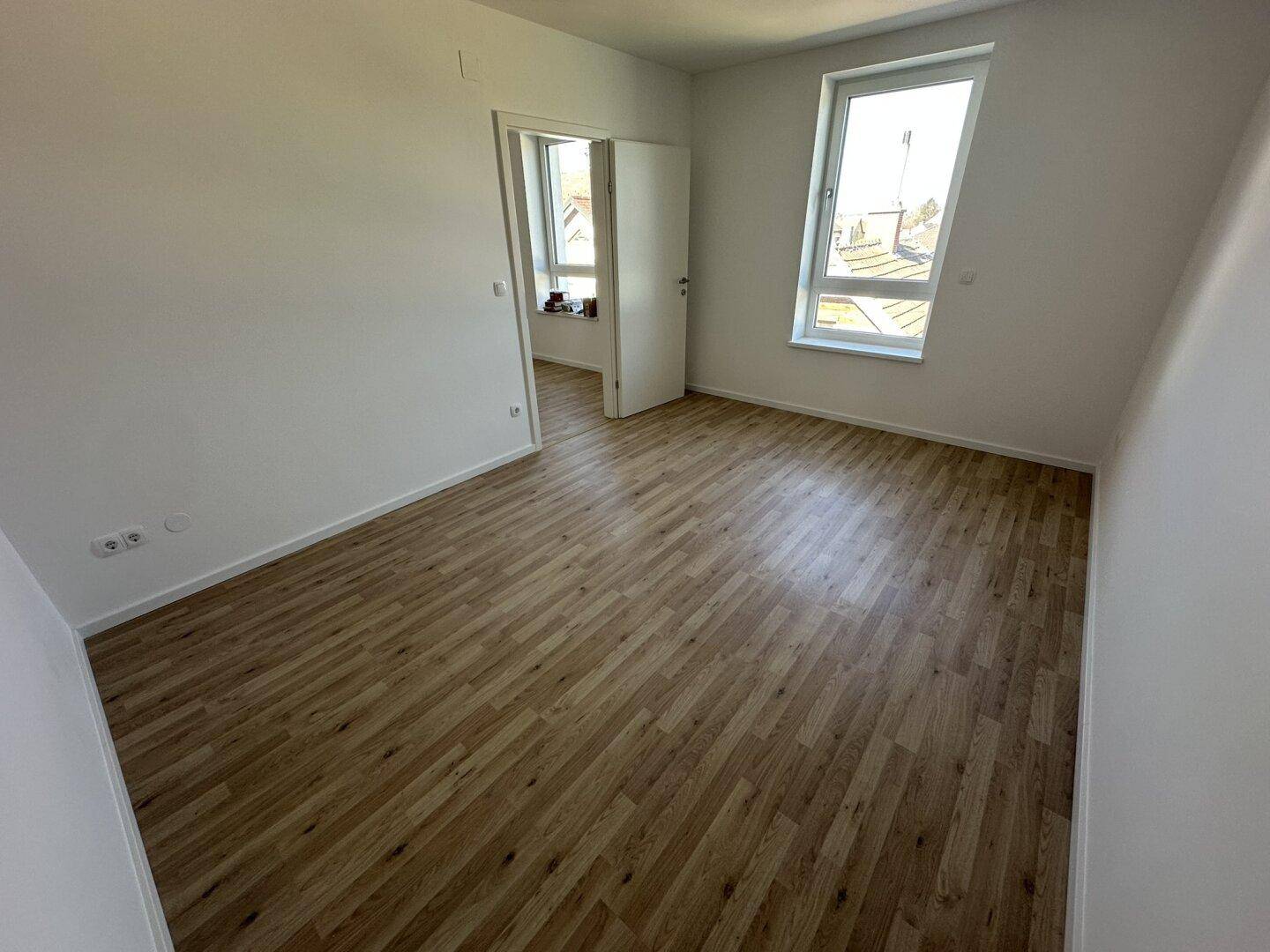 Lichtdurchfluteter Raum mit hellem Holzfußboden und zwei Fenstern, bereit zur individuellen Gestaltung.