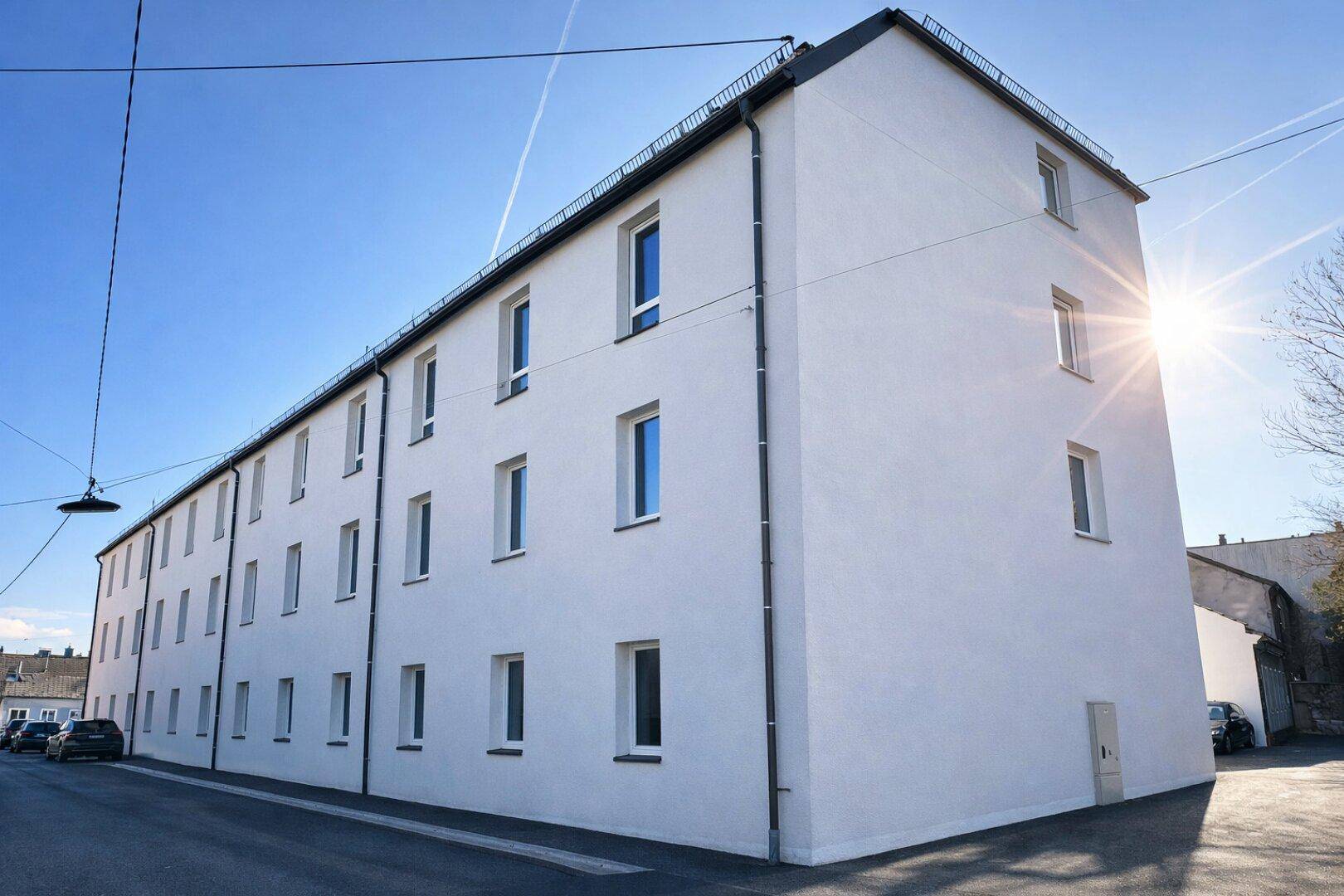 Langes, weißes Mehrfamilienhaus mit vielen Fenstern unter blauem Himmel und Sonnenschein.