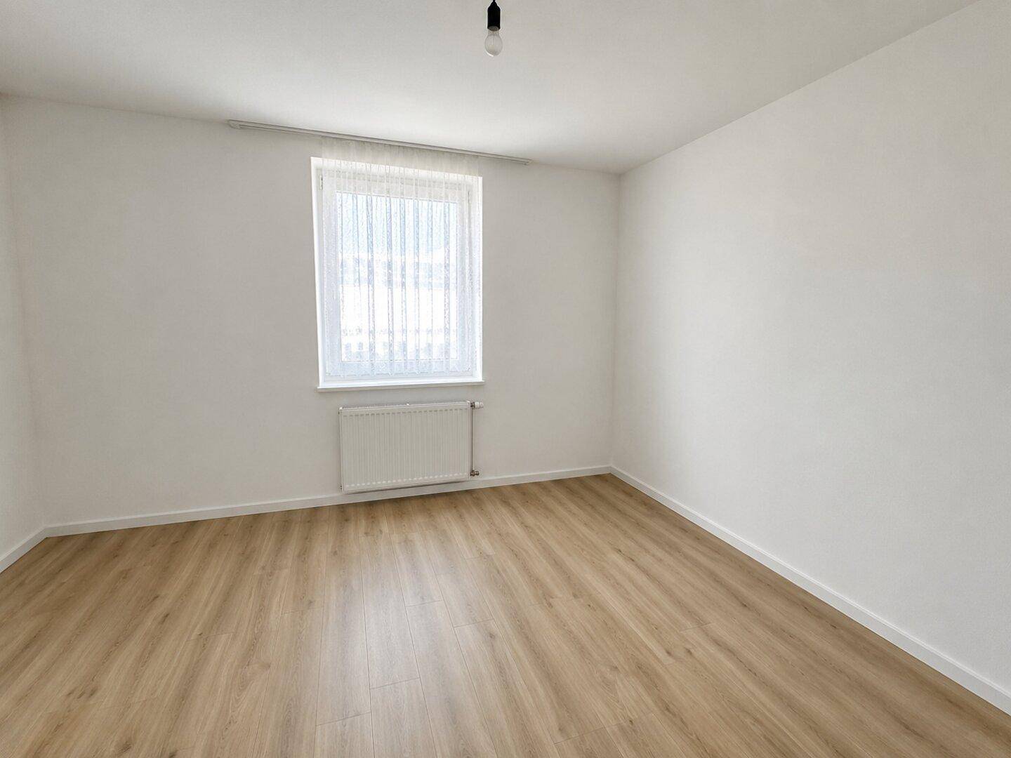 Unmöbliertes Zimmer mit Laminatboden, weiß gestrichenen Wänden und einem Heizkörper unter dem Fenster.