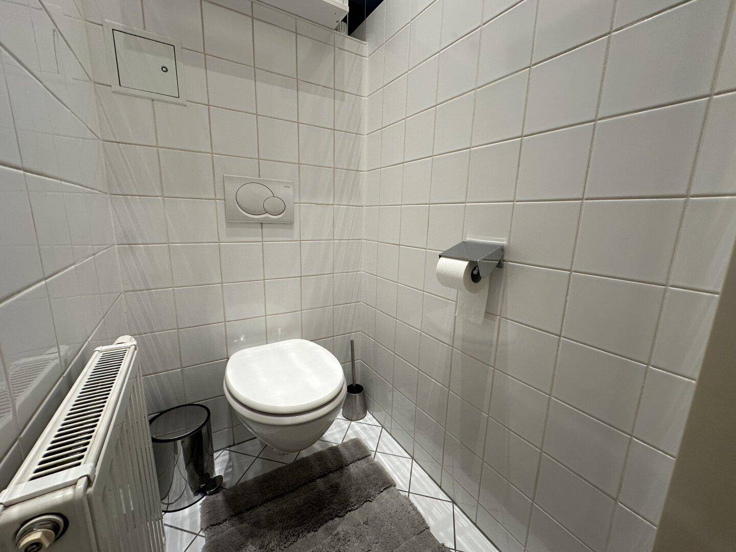 Separates WC mit weißen Wand- und Bodenfliesen und einem Heizkörper.