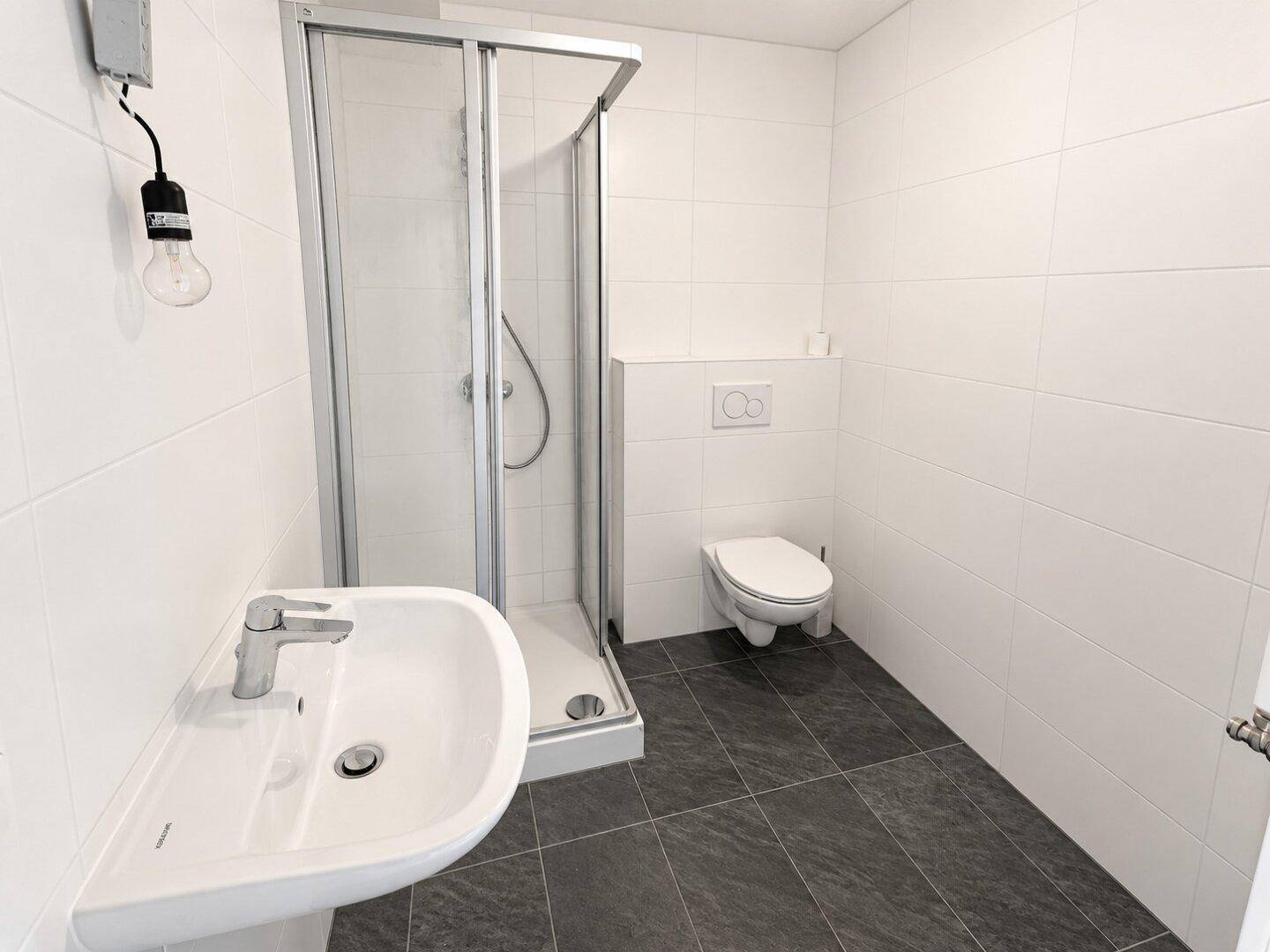 Modernes Badezimmer mit weißen Fliesen, dunklem Boden und einer Duschkabine.