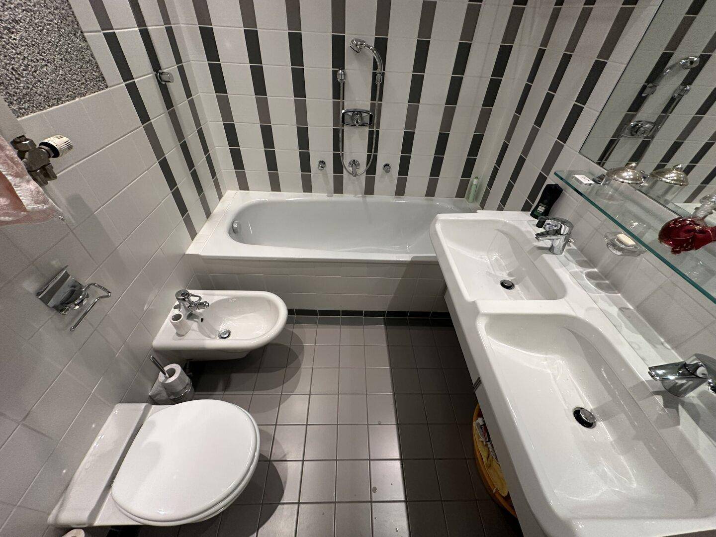 Funktionales Badezimmer mit Badewanne, Dusche, Bidet und Doppelwaschbecken. Die Wände sind mit markanten schwarz-weißen Streifenfliesen versehen.