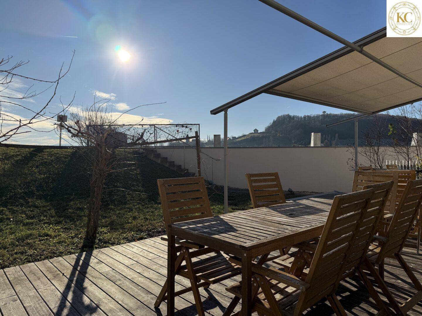 Gemütlicher Essbereich im Freien auf einer Holzterrasse mit Sonnenschutz und Blick auf die Hügellandschaft.