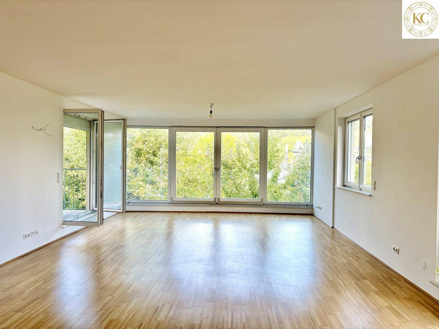 Helles, leeres Wohnzimmer mit Parkettboden und großen Fenstern, die viel Tageslicht hereinlassen.