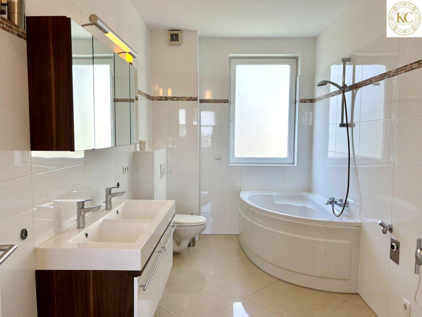 Modernes Badezimmer mit weißen Fliesen, Doppelwaschbecken, Eckbadewanne und Fenster für Tageslicht.