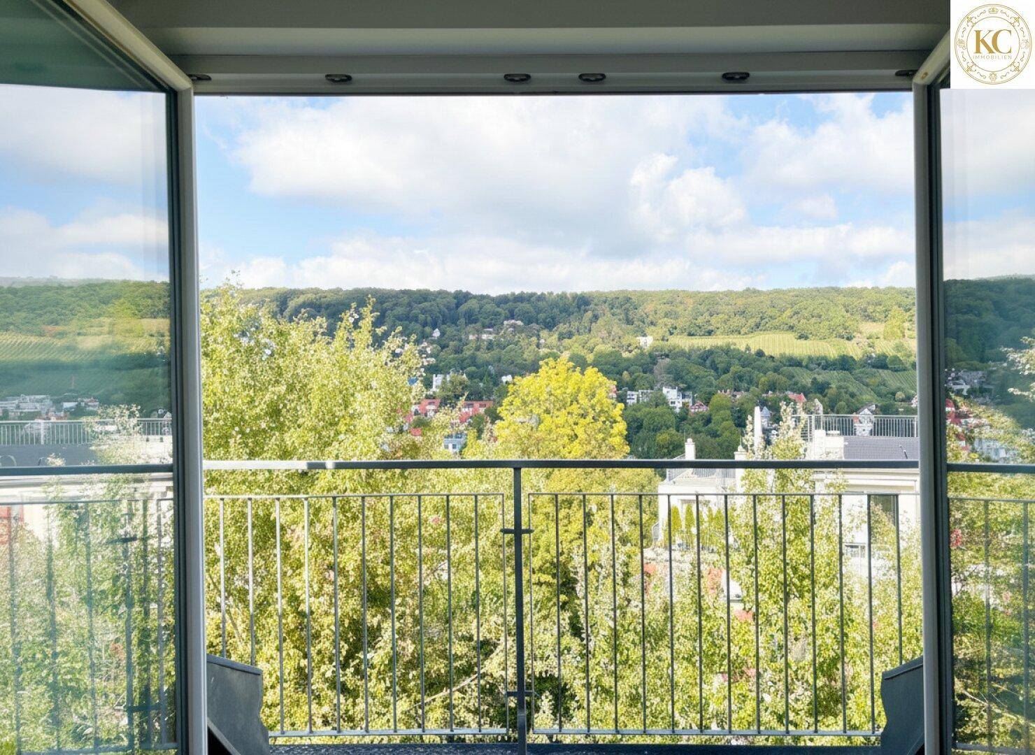 Blick vom Innenraum durch geöffnete Balkontüren auf die grüne Umgebung und den blauen Himmel.