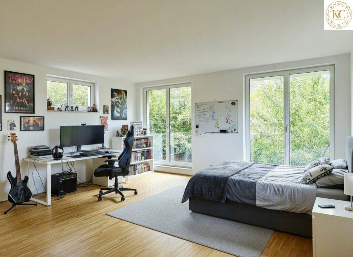 Helles Schlafzimmer mit Arbeitsbereich, Holzboden und großen Fenstern mit Blick ins Grüne.
