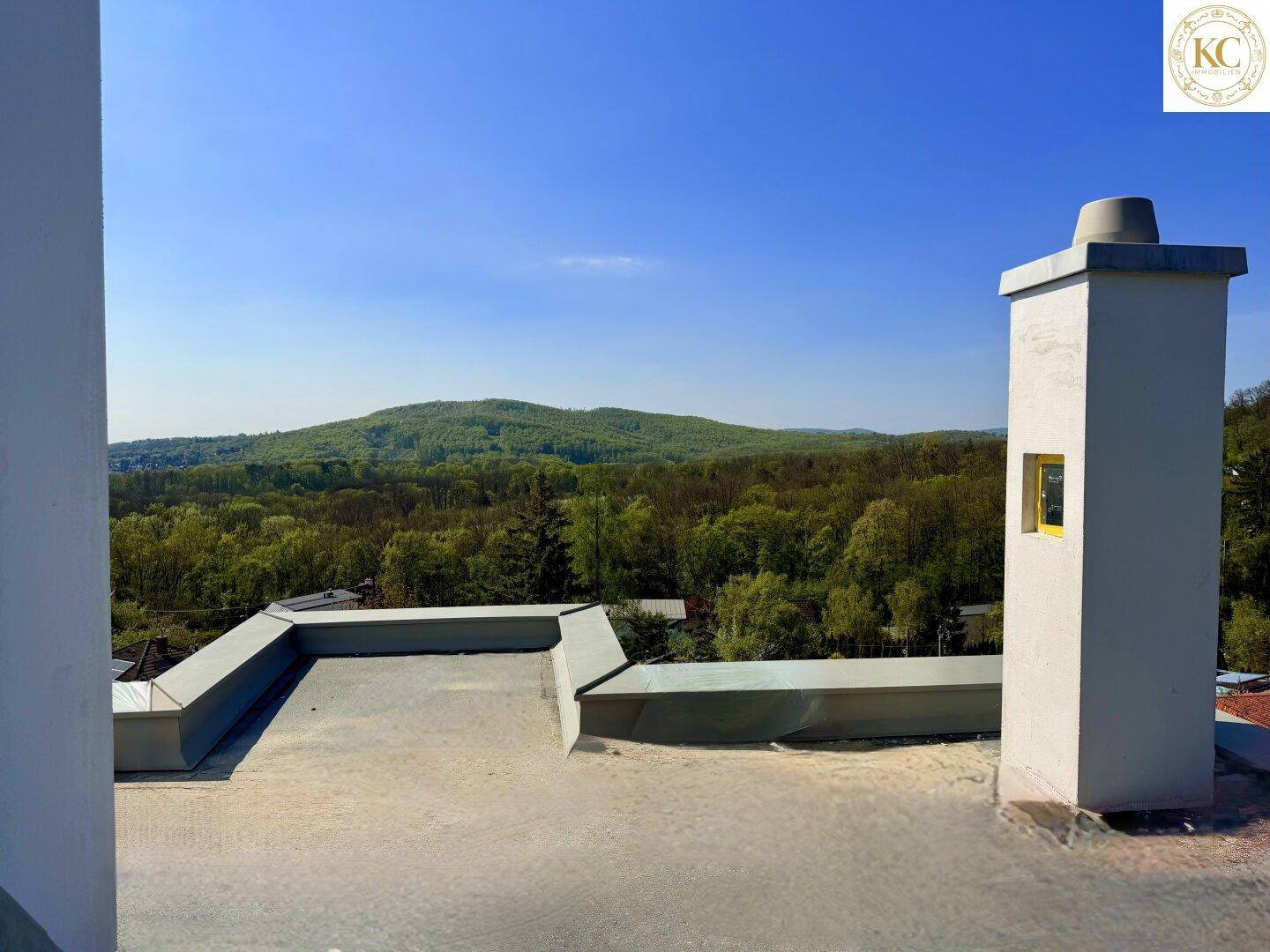 Blick von einem Flachdach auf eine weite grüne Hügellandschaft unter blauem Himmel.