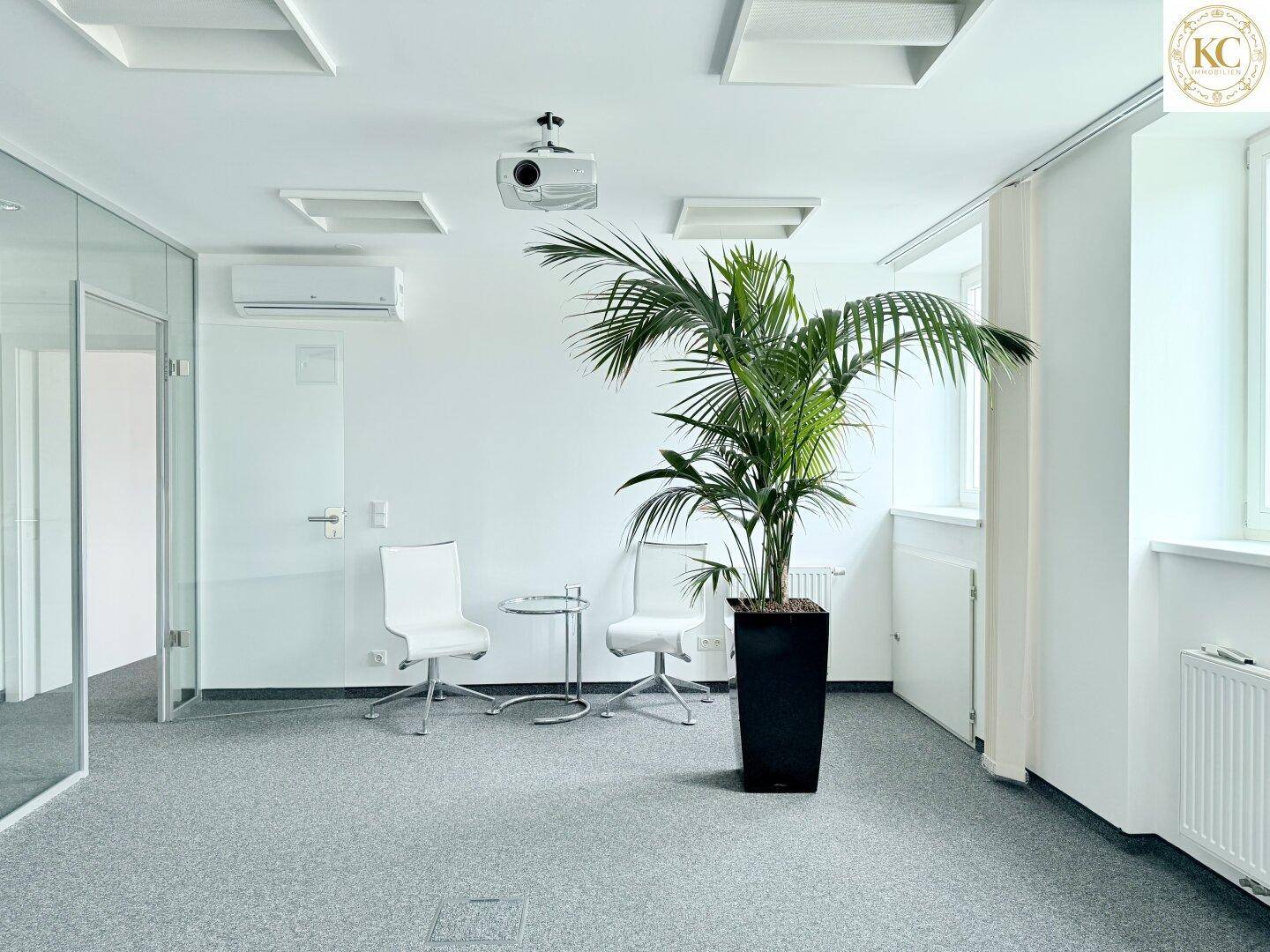 Helles Büro mit Teppichboden, Glaswänden, Klimaanlage und einem Projektor an der Decke.