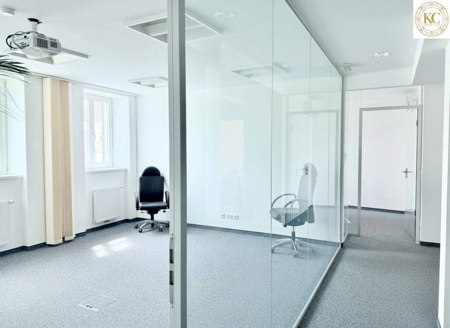 Büro mit Teppichboden, Glaswänden und Blick auf den Flur sowie Tageslicht von außen.