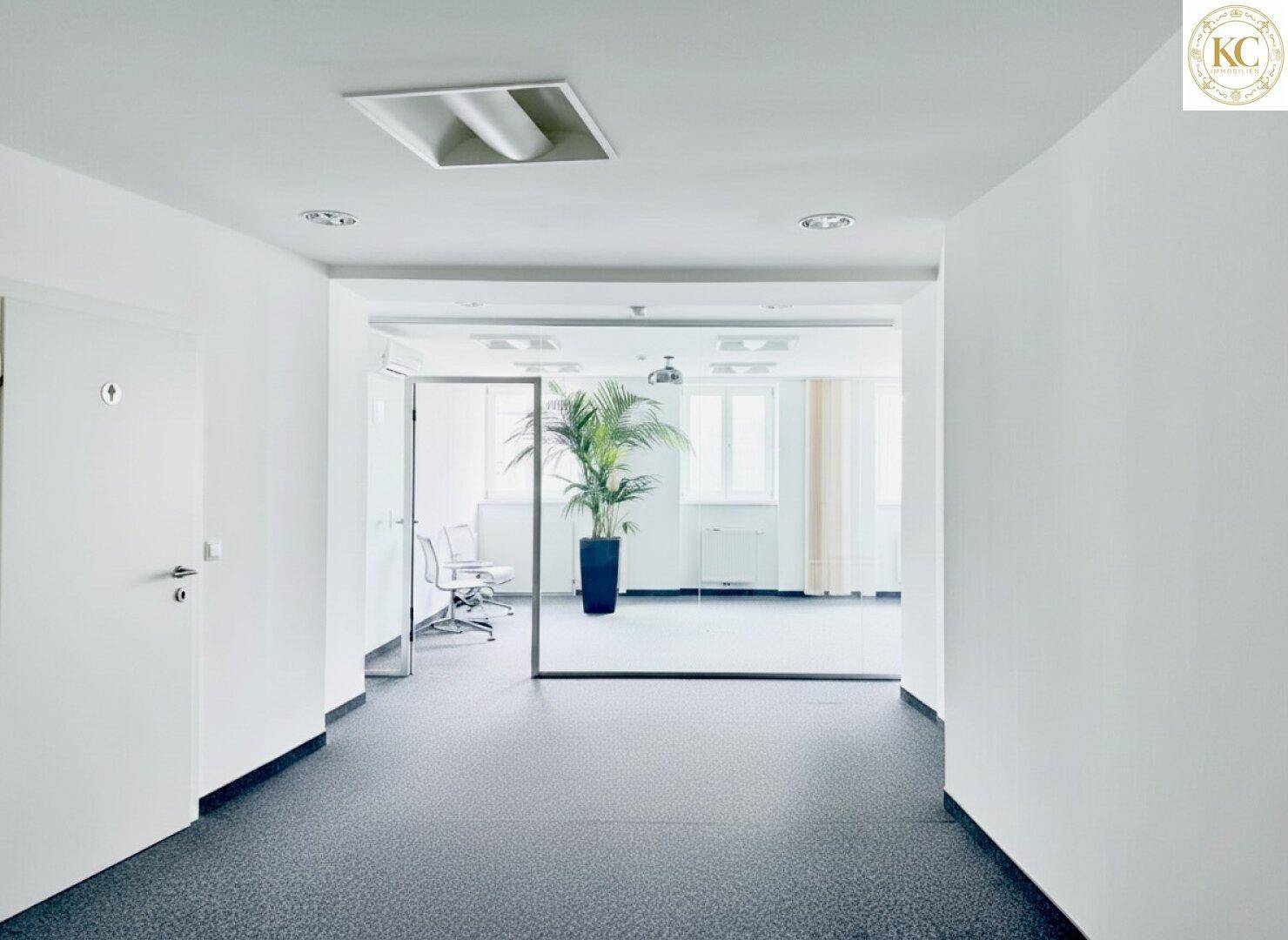 Heller Flur mit dunklem Teppichboden und Zugang zu weiteren Büroräumen mit Glaswänden.