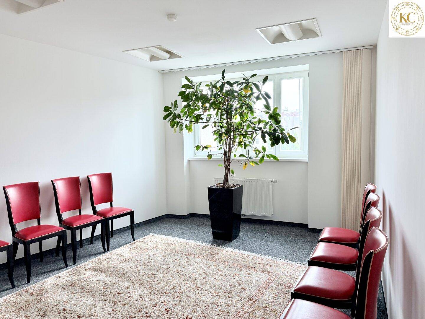 Wartebereich oder Besprechungsraum mit roten Stühlen, Teppichboden und einem großen Fenster.
