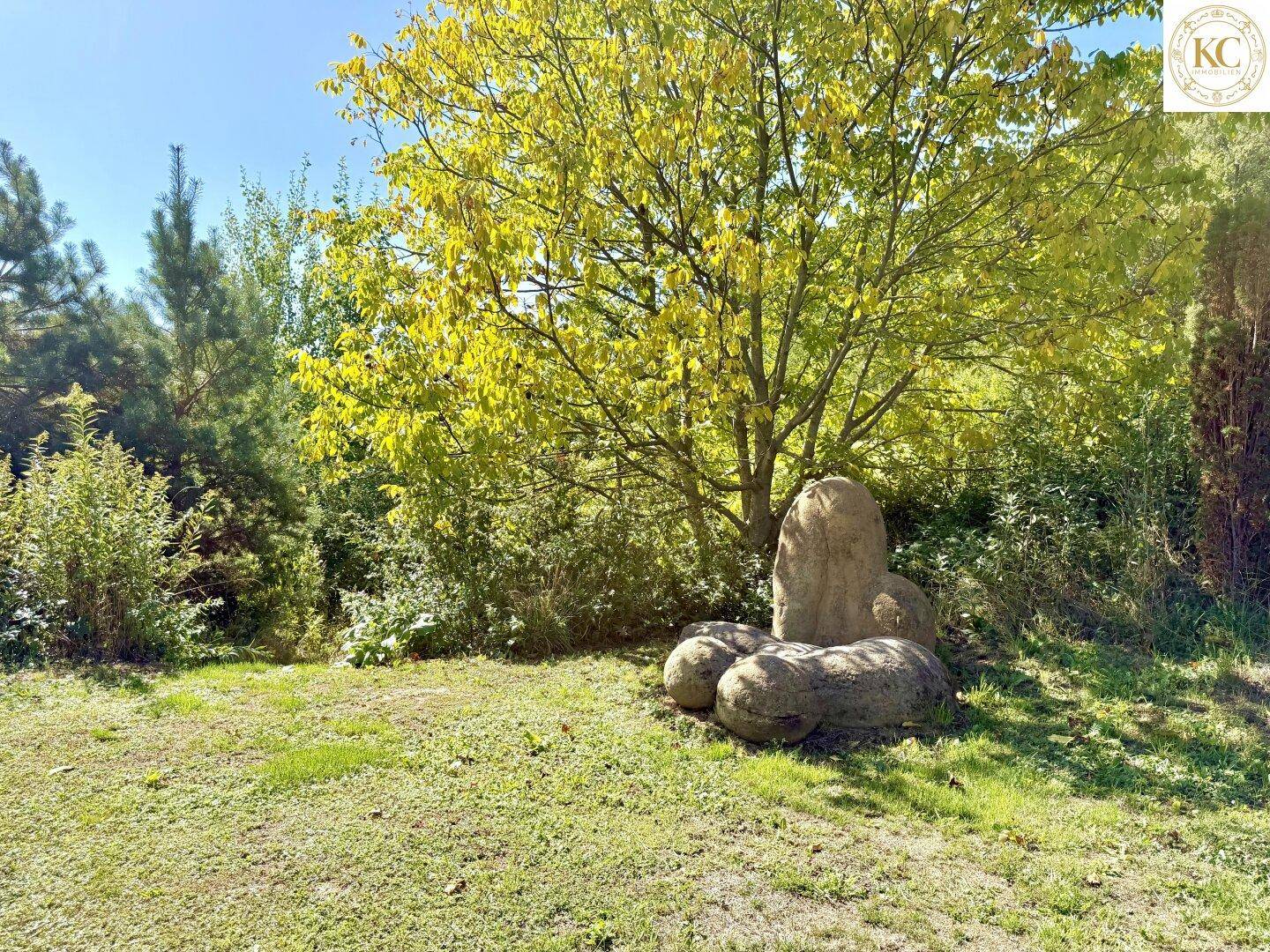 Natürlicher Gartenbereich mit Rasenfläche, Bäumen und einer markanten Felsformation unter blauem Himmel.