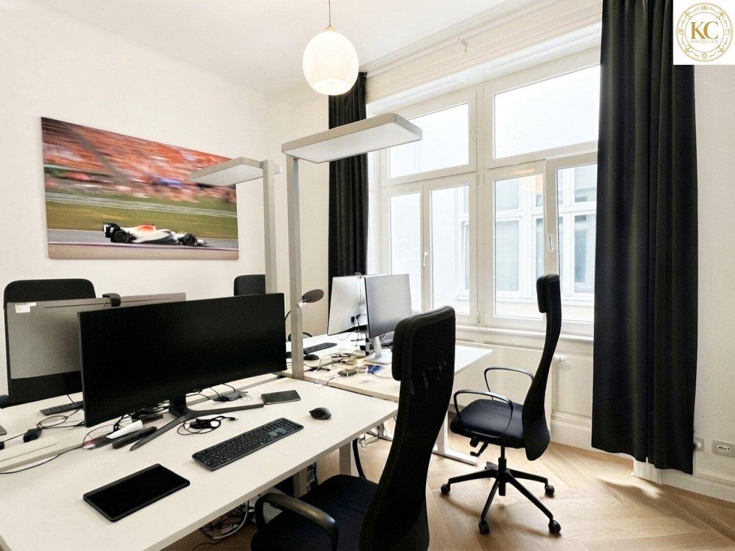 Geräumiges Büro mit mehreren Arbeitsplätzen, großen Fenstern und dunklen Vorhängen.