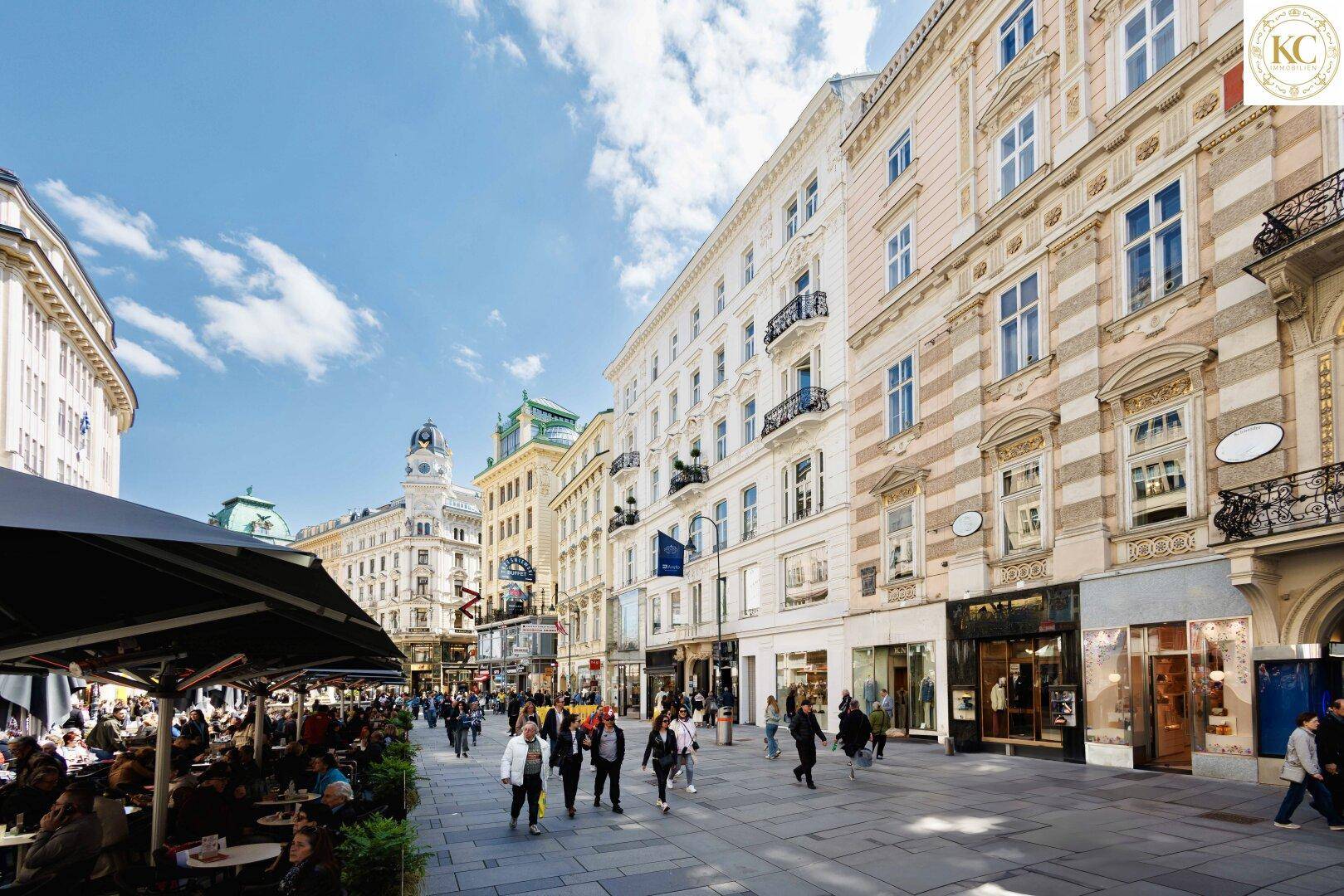 Belebte Einkaufsstraße mit historischen Gebäuden, Geschäften und Außengastronomie unter blauem Himmel.