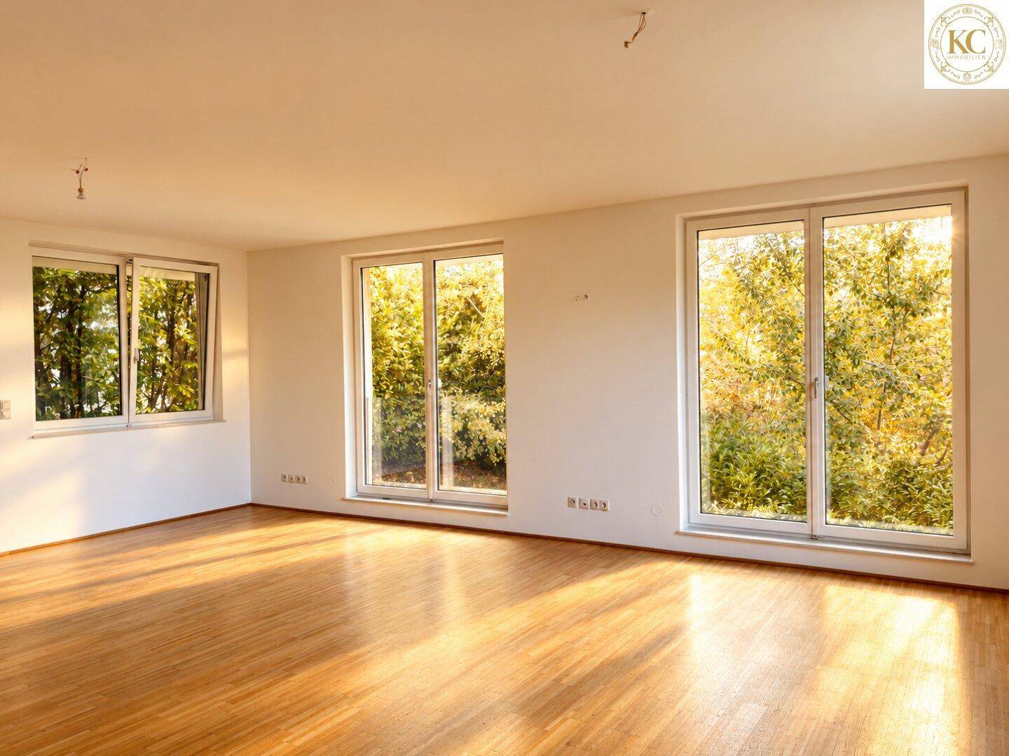 Helles, leeres Wohnzimmer mit Parkettboden und großen Fenstern mit Blick auf die grüne Umgebung.