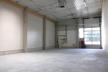 Großer, leerer Industrieraum mit hohen Decken, Betonboden und einem großen Rolltor mit Glasfenstern.