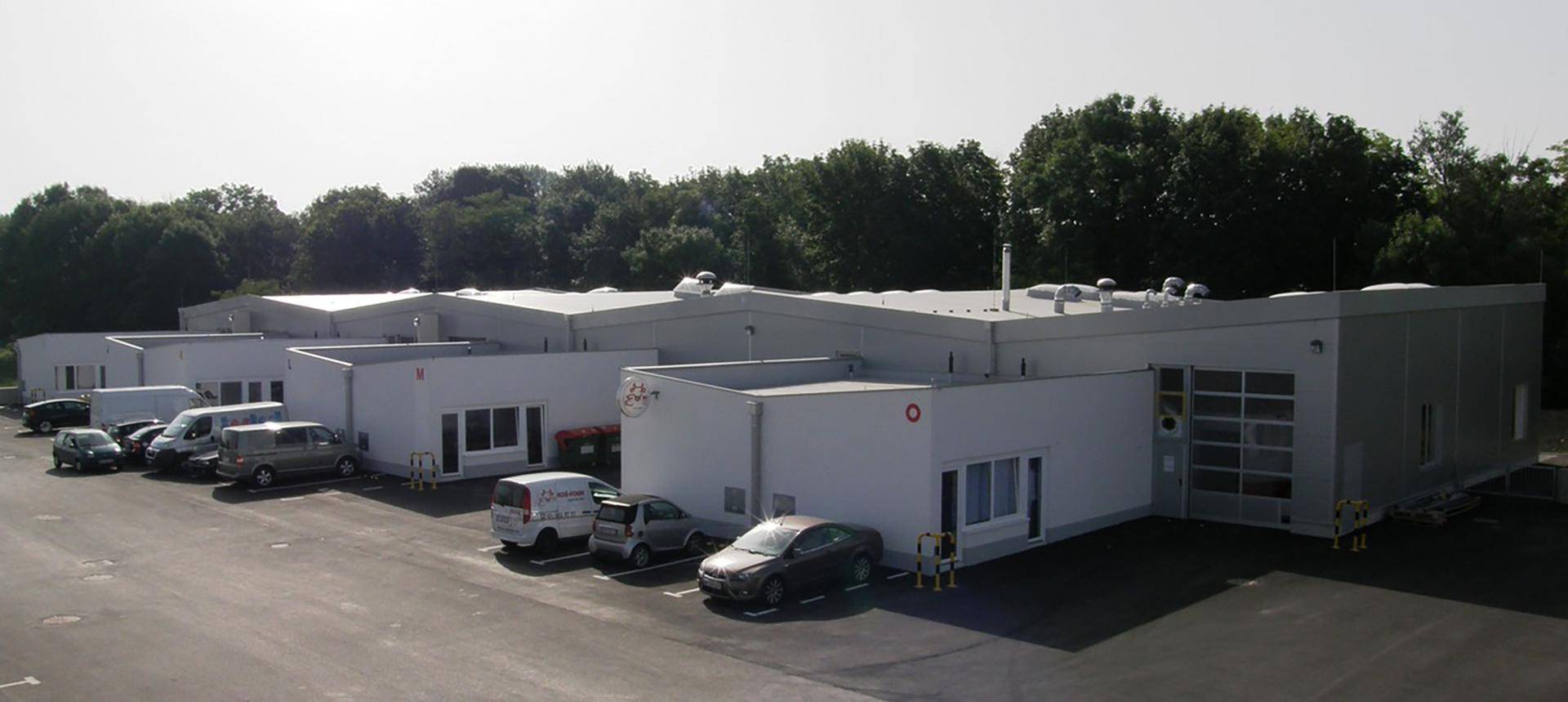Moderne Gewerbegebäude mit klaren Linien und großem Parkplatz, umgeben von grünen Bäumen.