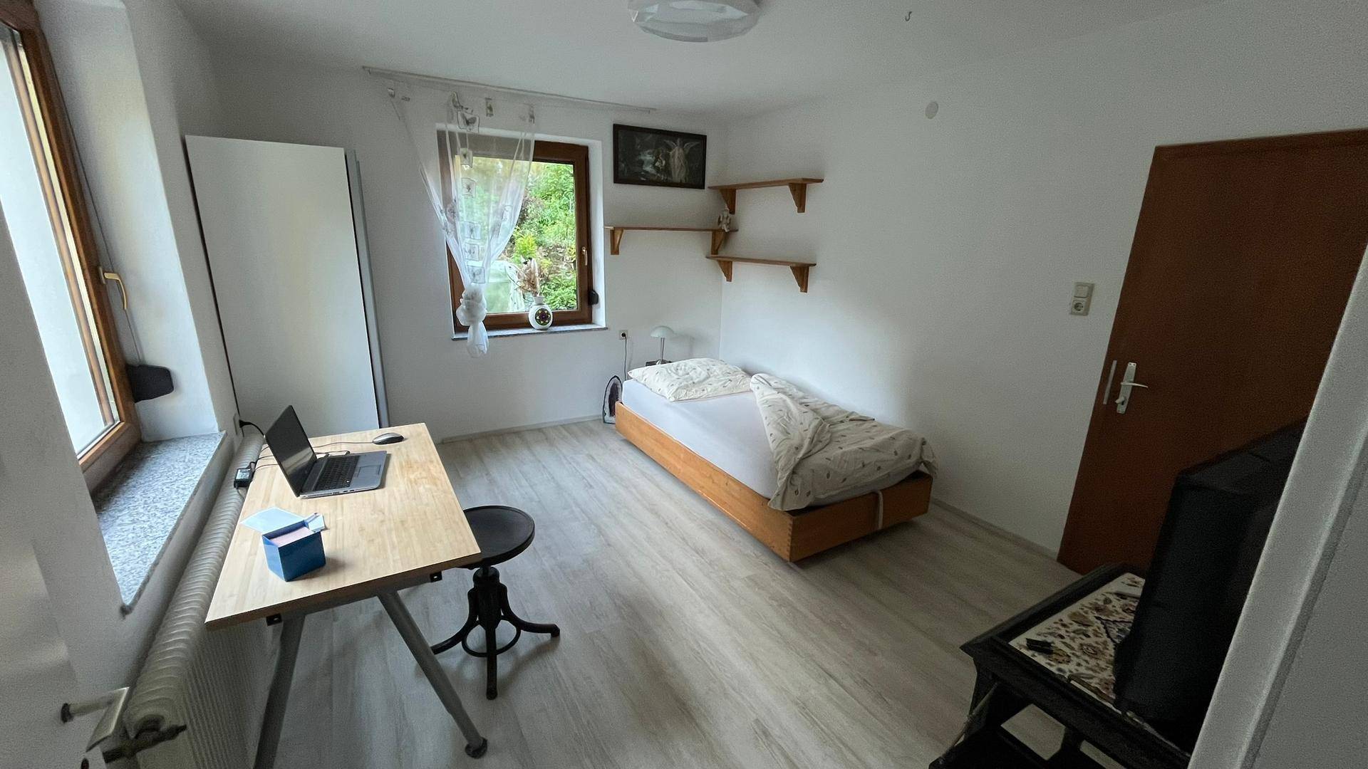 Freundliches Schlafzimmer mit Arbeitsbereich, Einzelbett und Blick in die Natur durch die Fenster.