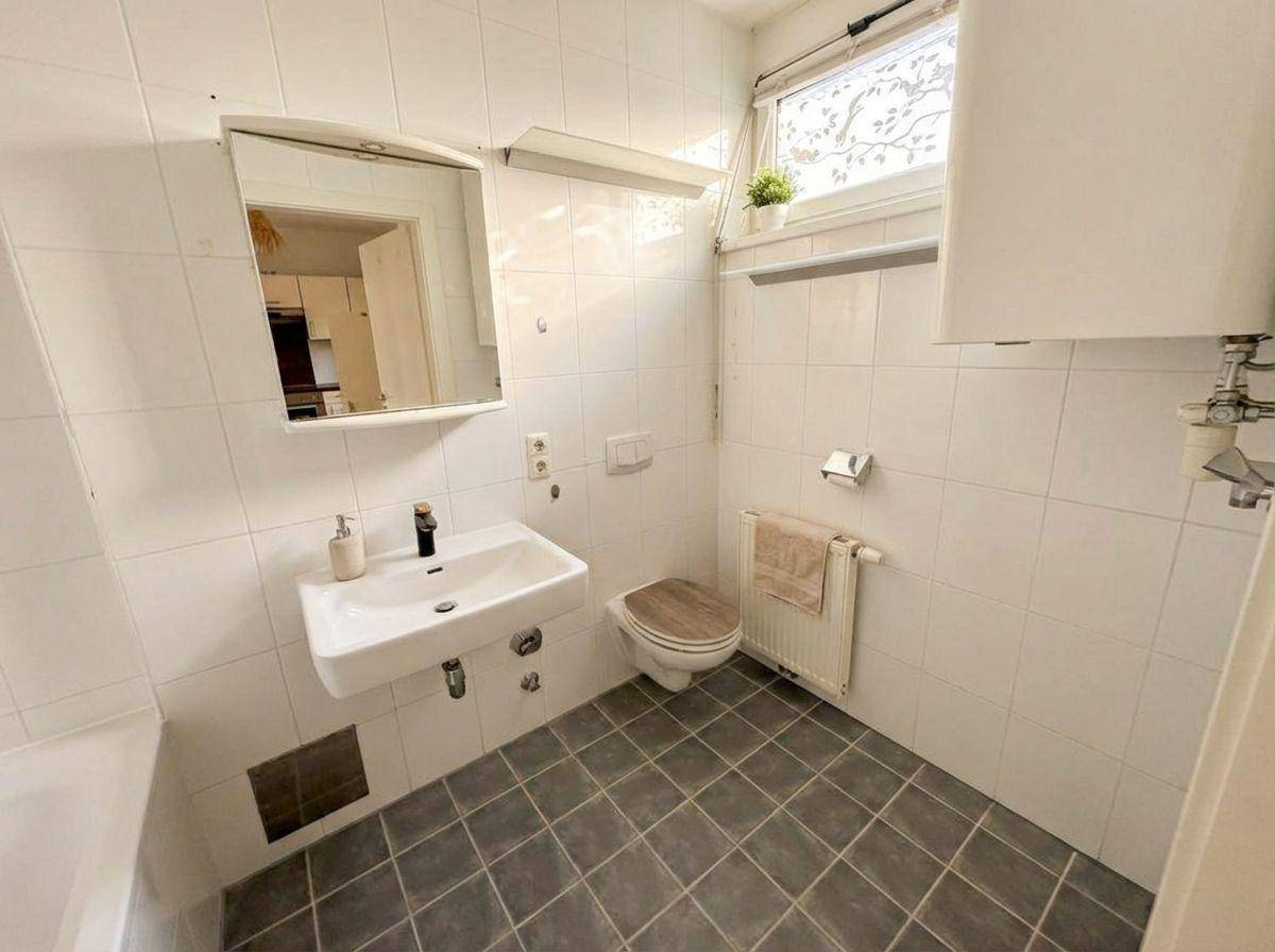 Helles Badezimmer mit weißen Fliesen, Fenster und einer Badewanne mit Duschfunktion.