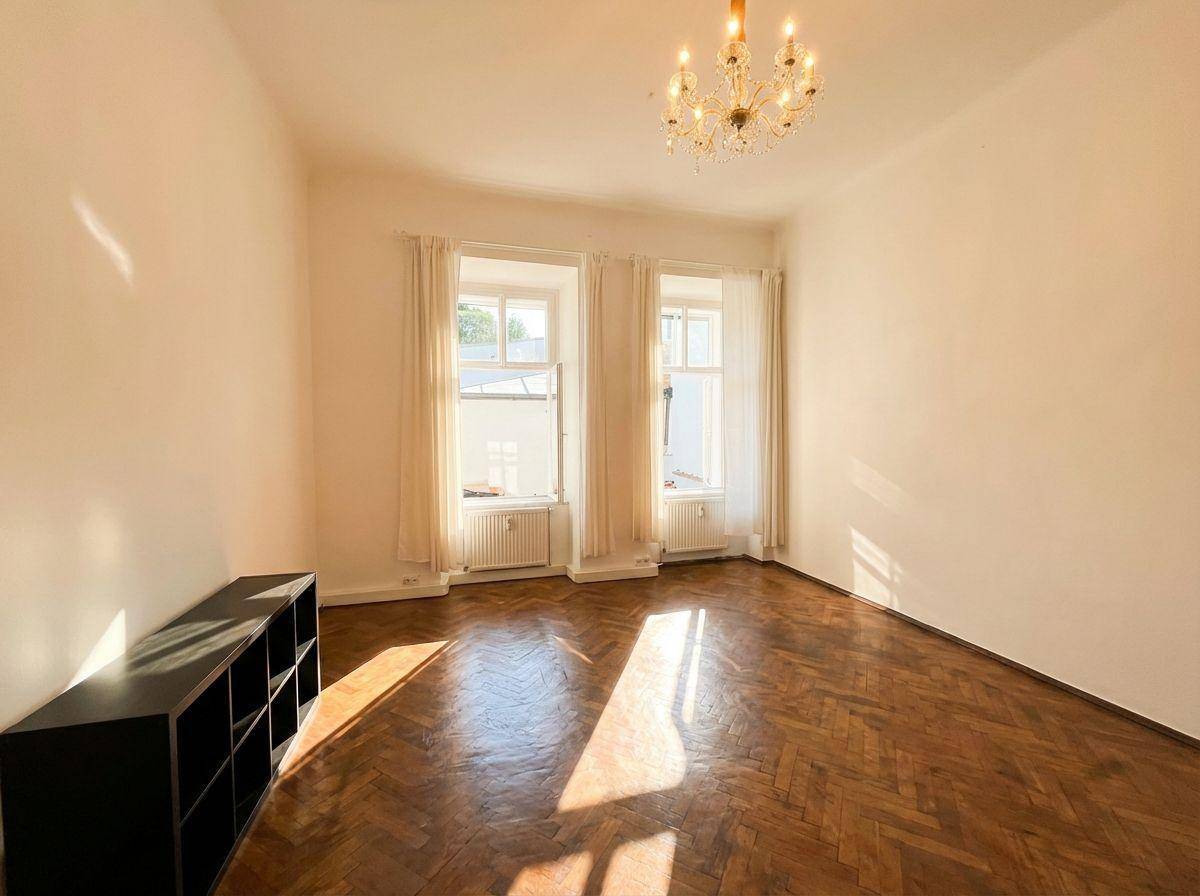 Helles Zimmer mit Fischgrätparkett, zwei Fenstern und einem Kronleuchter an der Decke.