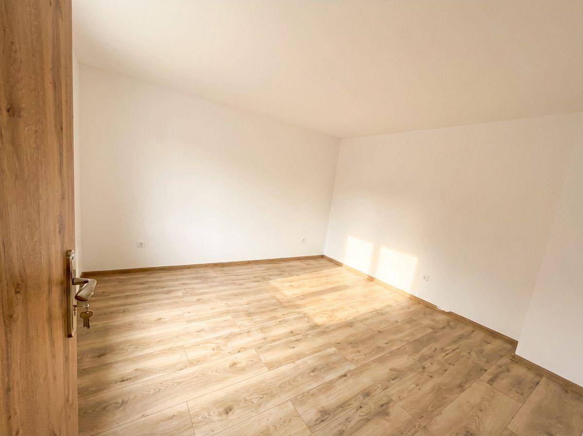 Helles, unmöbliertes Zimmer mit Laminatboden und einer Holztür, durchflutet von Sonnenlicht.