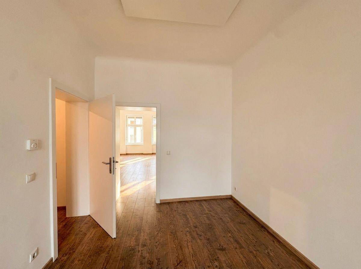 Heller Raum mit Holzboden und offener Tür zu einem weiteren Zimmer, das viel Licht empfängt.