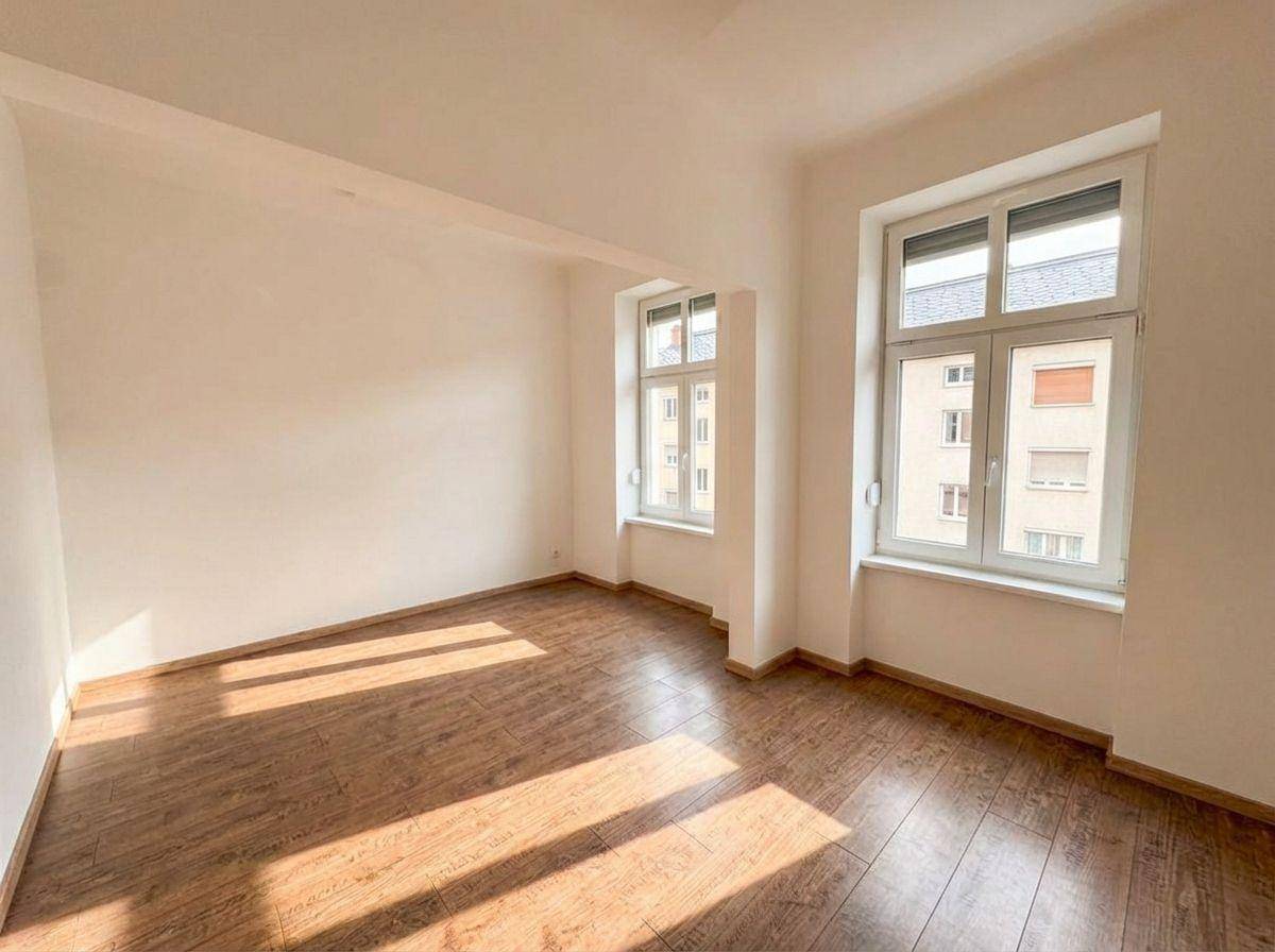Geräumiges Wohnzimmer mit Holzboden und großen Fenstern, die den Raum mit Sonnenlicht füllen.