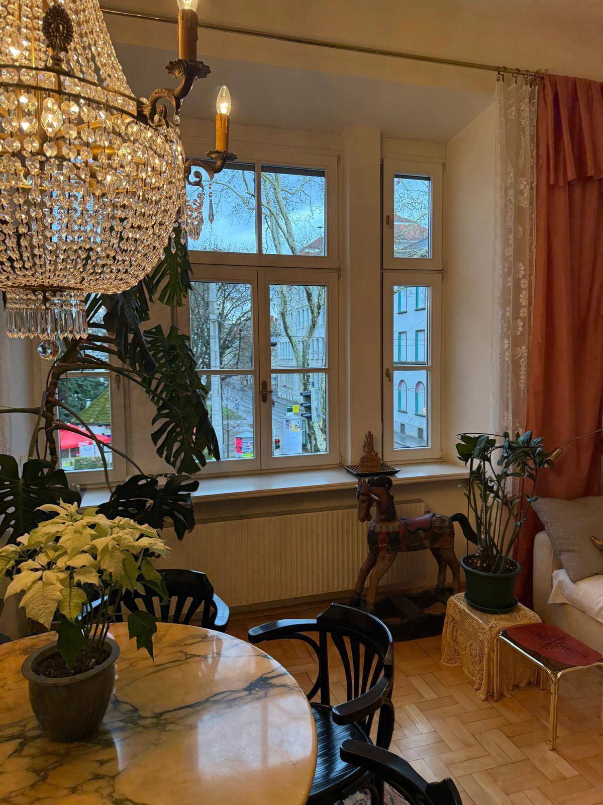 Heller Wohn-Essbereich mit großem Fenster, Kronleuchter und Blick auf die Stadt.