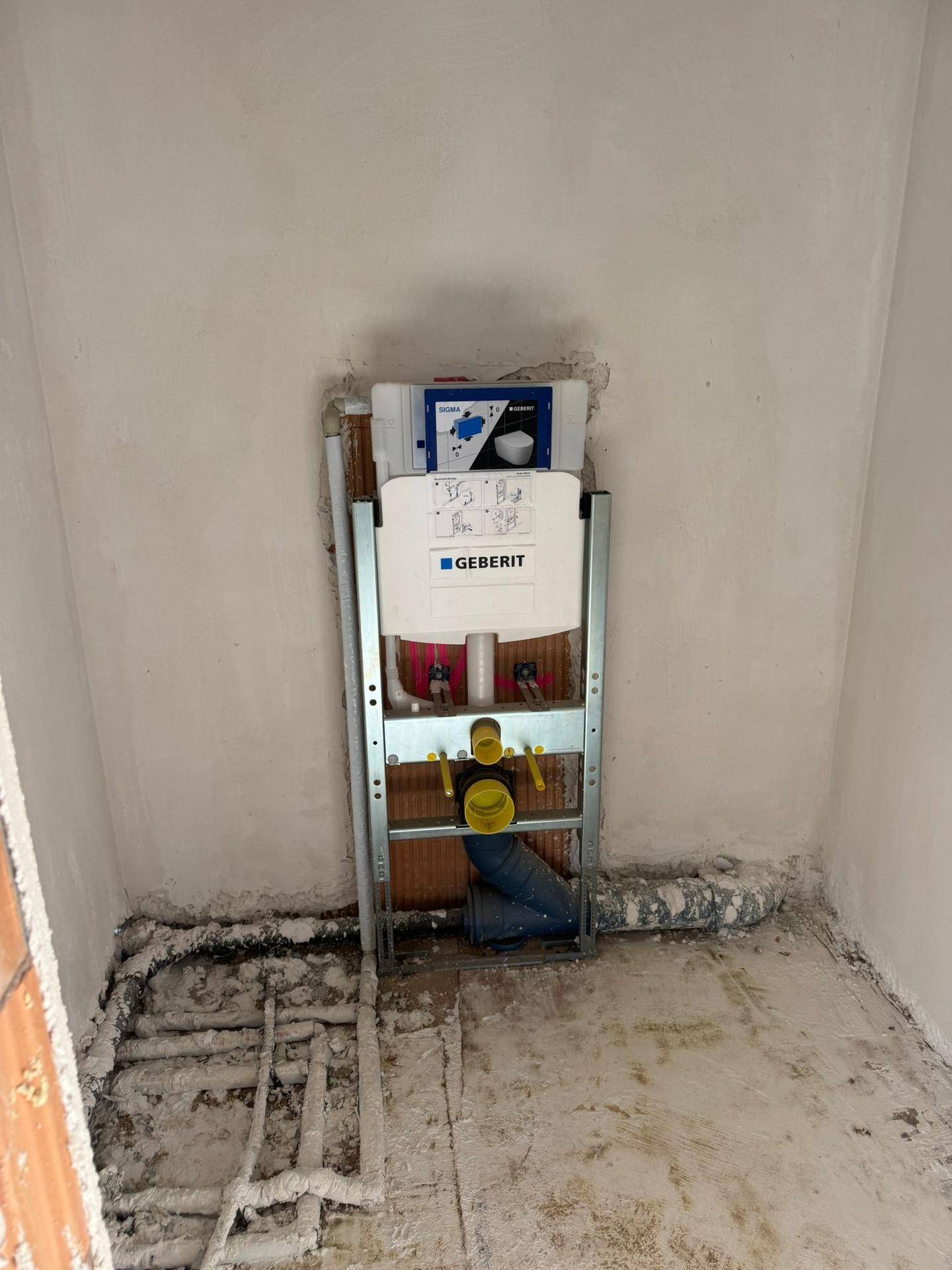 Badezimmer im Rohbau mit sichtbarer Vorinstallation für eine Wandtoilette und Leitungen am Boden.