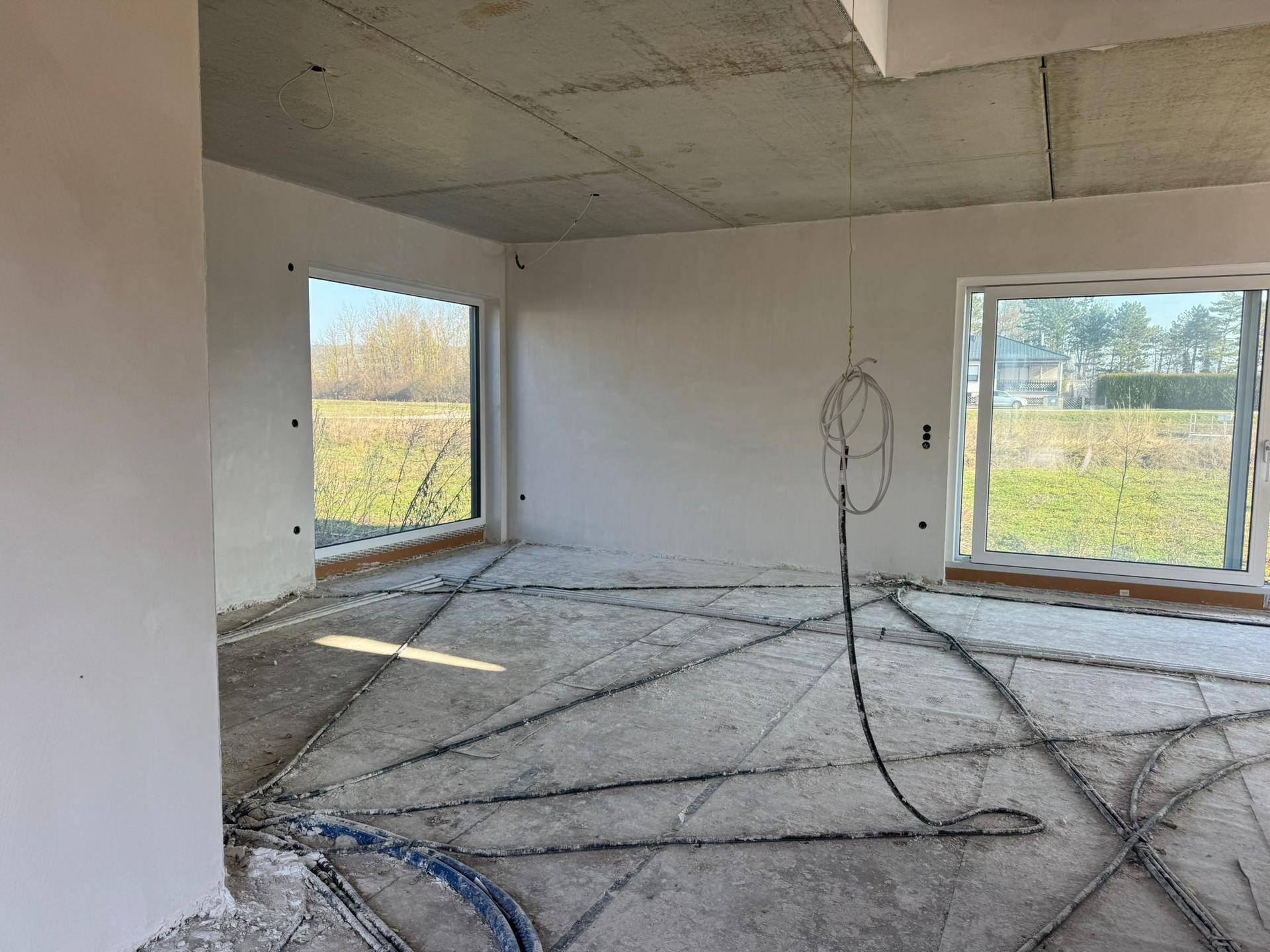 Heller Innenraum im Rohbau mit großen Fenstern und sichtbaren Leitungen für die Fußbodenheizung.