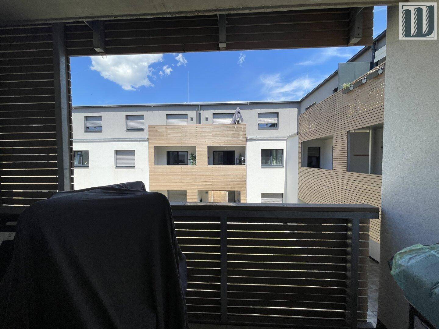 Blick vom Balkon auf den begrünten Innenhof des Wohnkomplexes unter blauem Himmel.