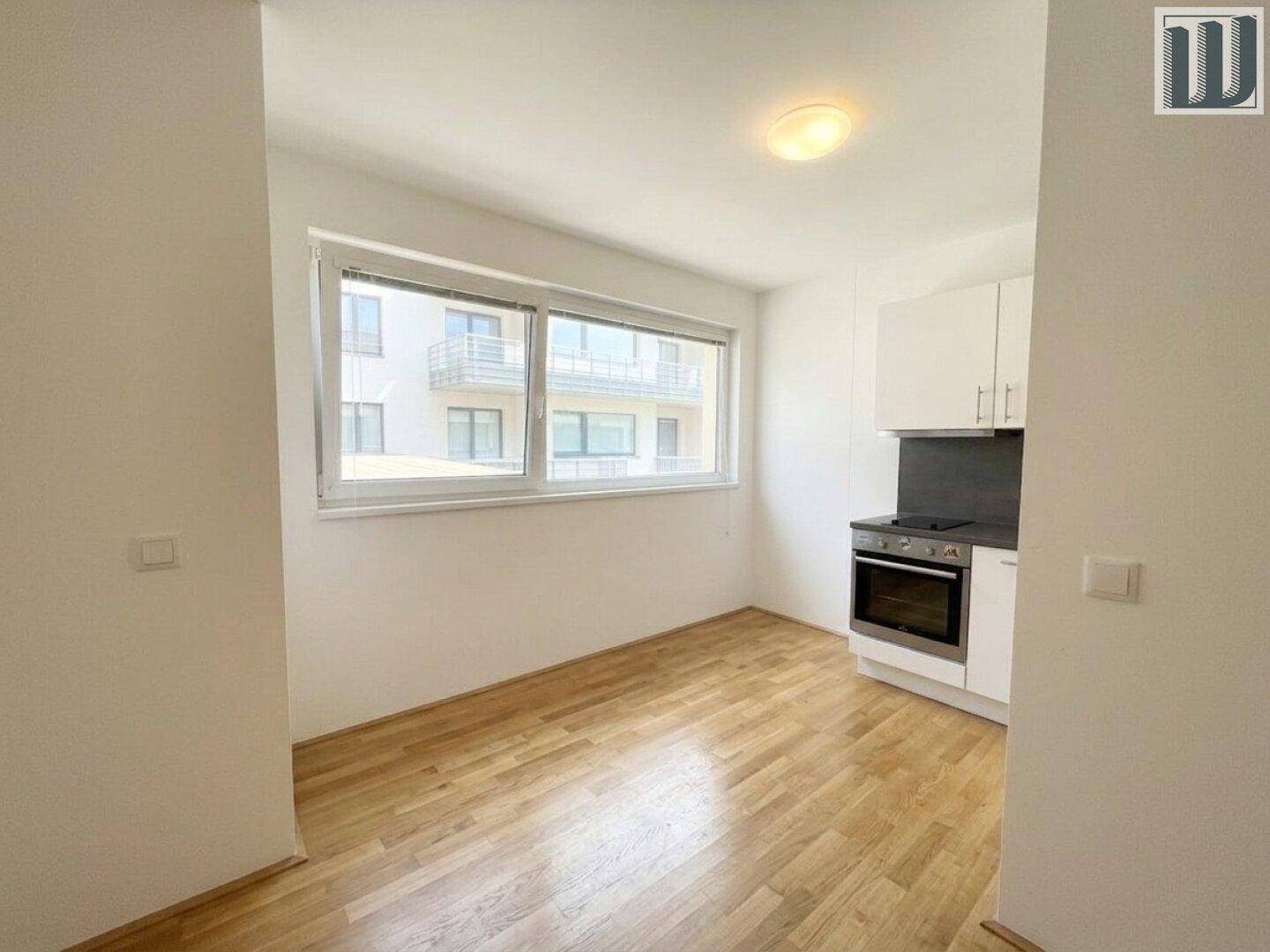 Küchenbereich mit Parkettboden, Fenster und einfacher Einbauküche in einem hellen Raum.