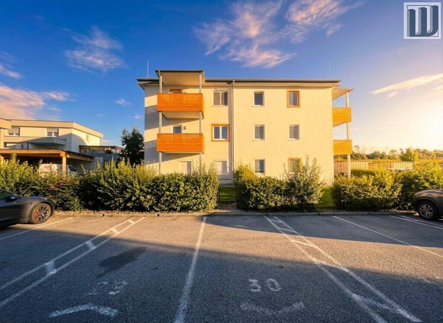 Neubau mit Balkonen und Parkplätzen bei Sonnenuntergang, umgeben von grüner Landschaft.