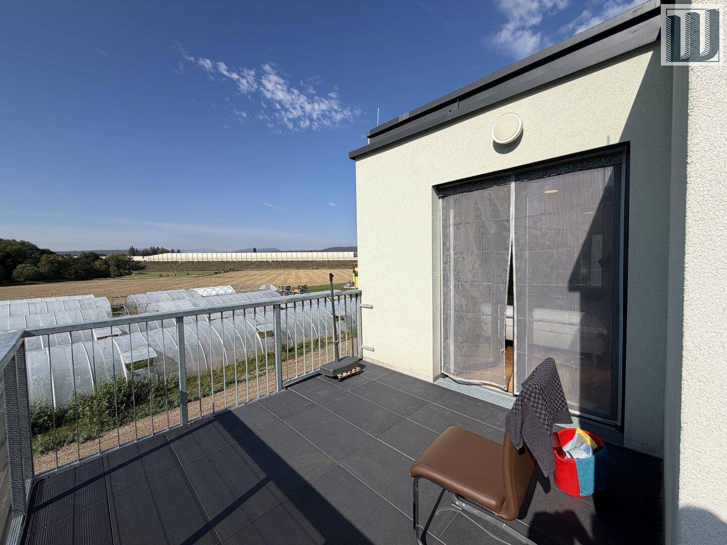 Geräumiger Balkon mit dunklem Bodenbelag und Ausblick auf Felder und Gewächshäuser.