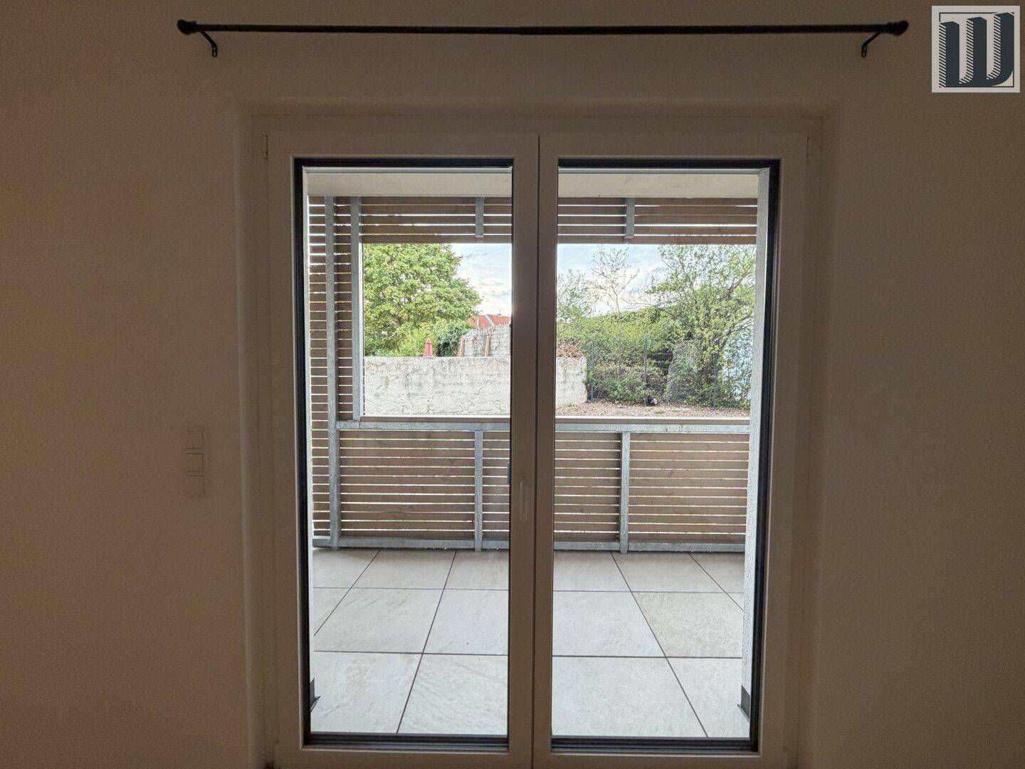 Blick durch die Balkontür auf einen gefliesten Balkon mit Holzverkleidung und grüner Umgebung.