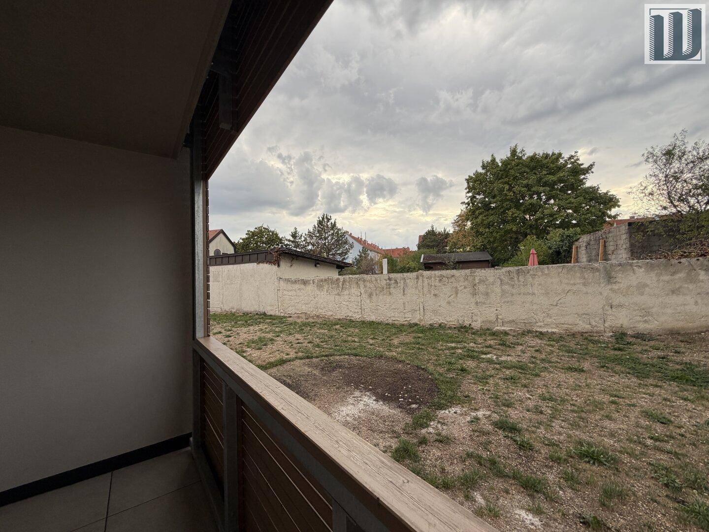 Aussicht vom Balkon auf eine Rasenfläche, umgeben von einer Mauer und Bäumen unter bewölktem Himmel.