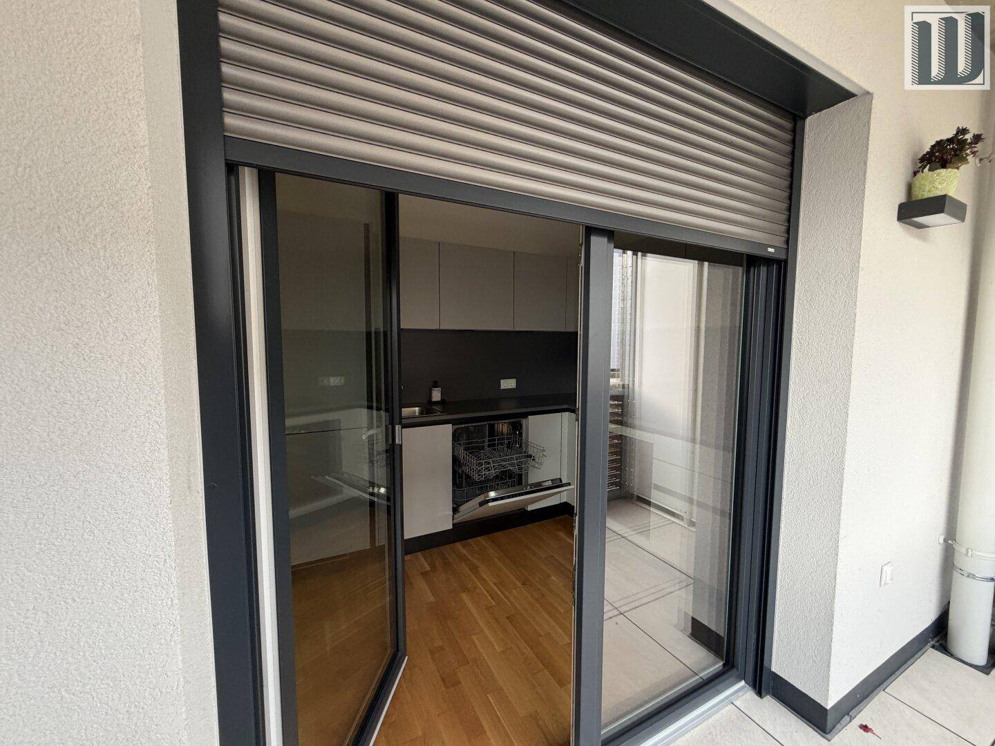 Blick vom Balkon in die Küche durch eine große Glasschiebetür mit Rollladen.