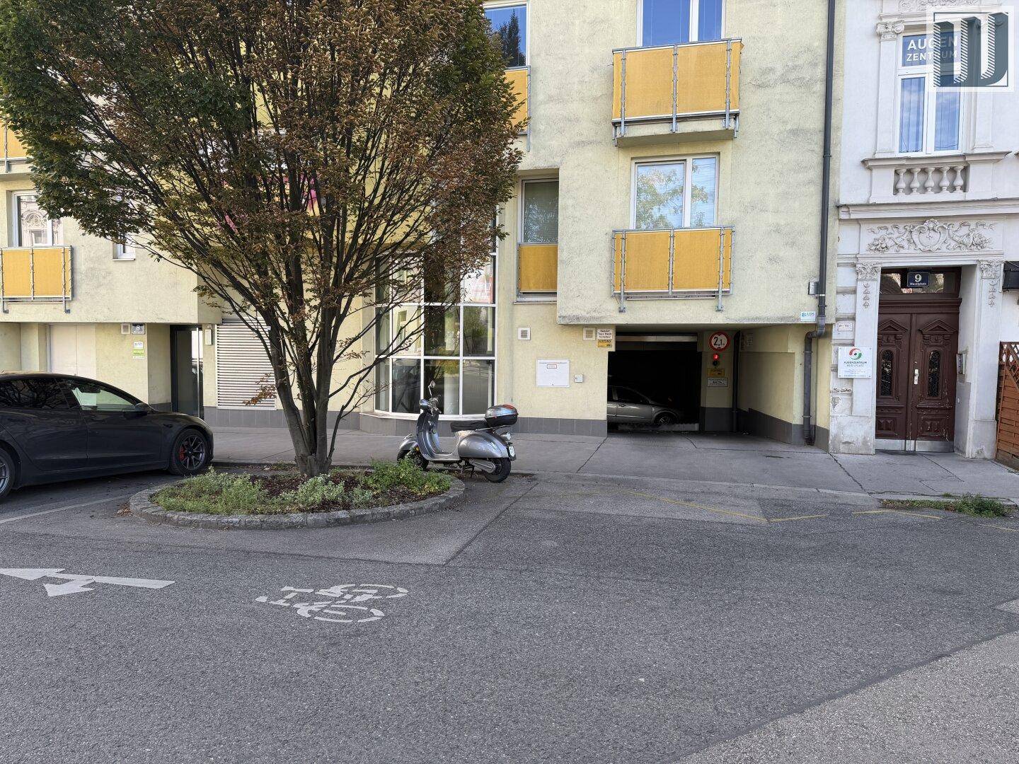 Außenansicht eines Wohngebäudes mit Balkonen, einem Baum und der Einfahrt zur Tiefgarage.