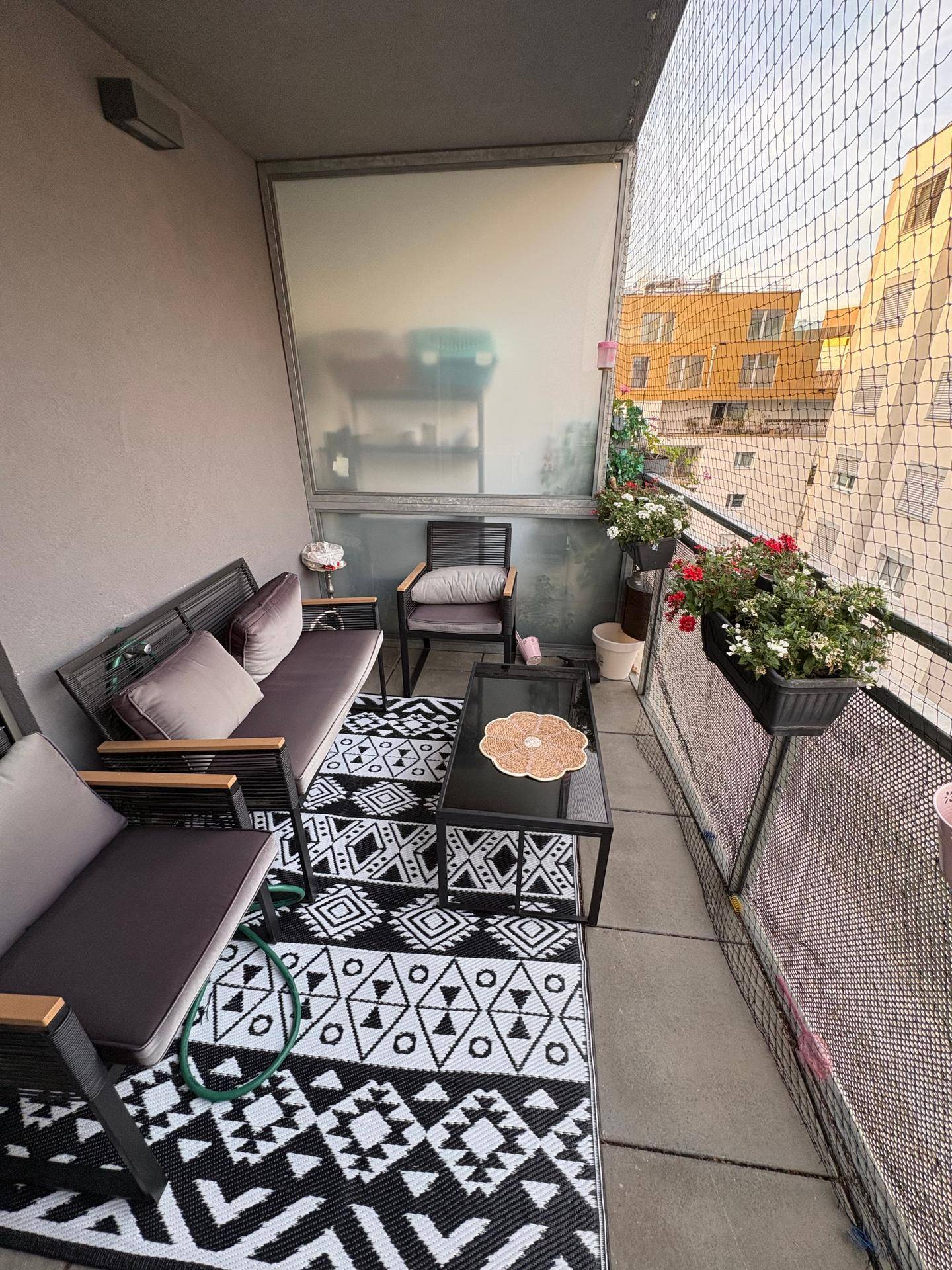Gemütlicher Balkon mit Outdoor-Möbeln, Teppich und Blick auf umliegende Gebäude.
