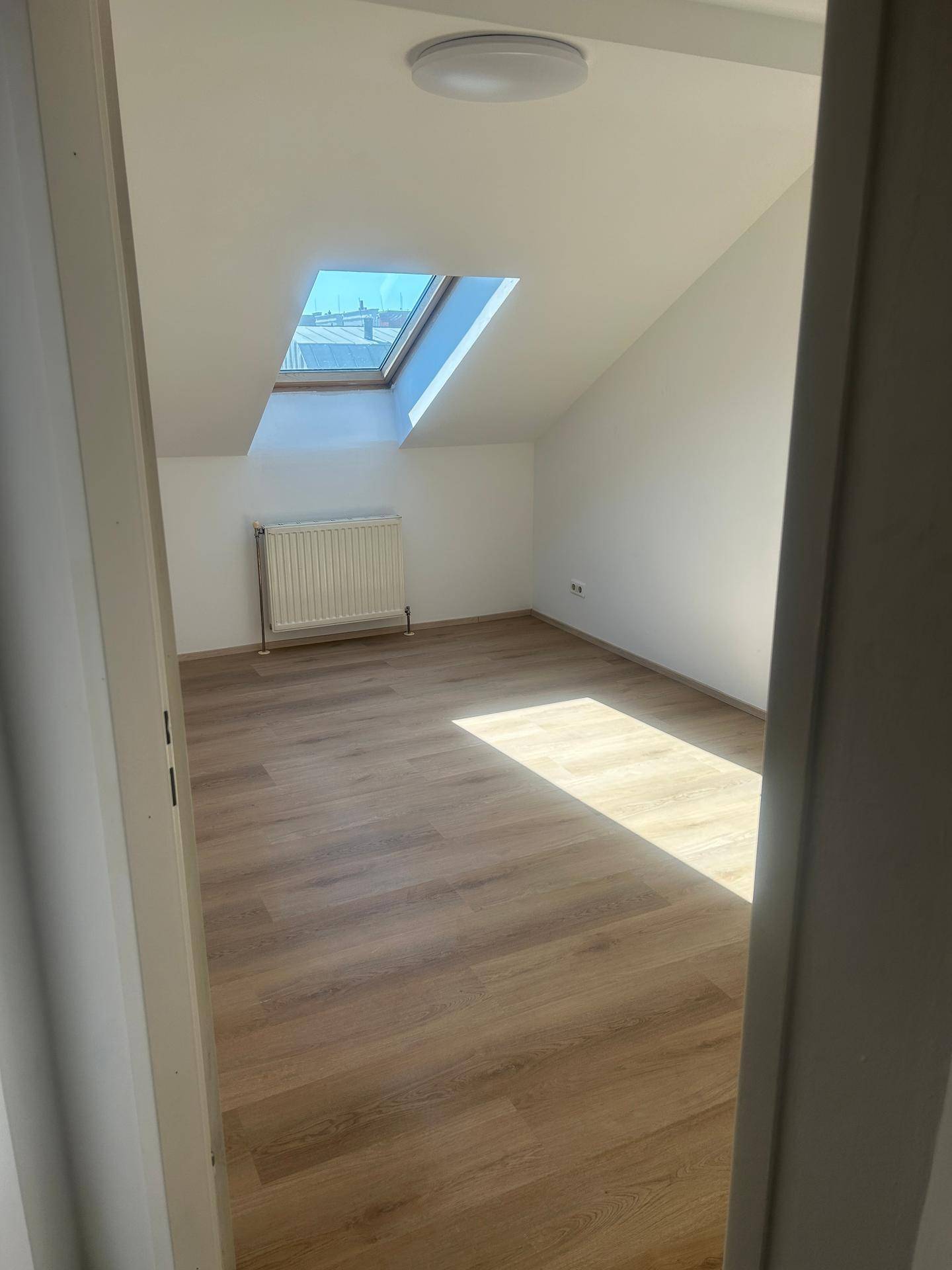 Helles Zimmer im Dachgeschoss mit Laminatboden, Heizkörper und großem Dachfenster.
