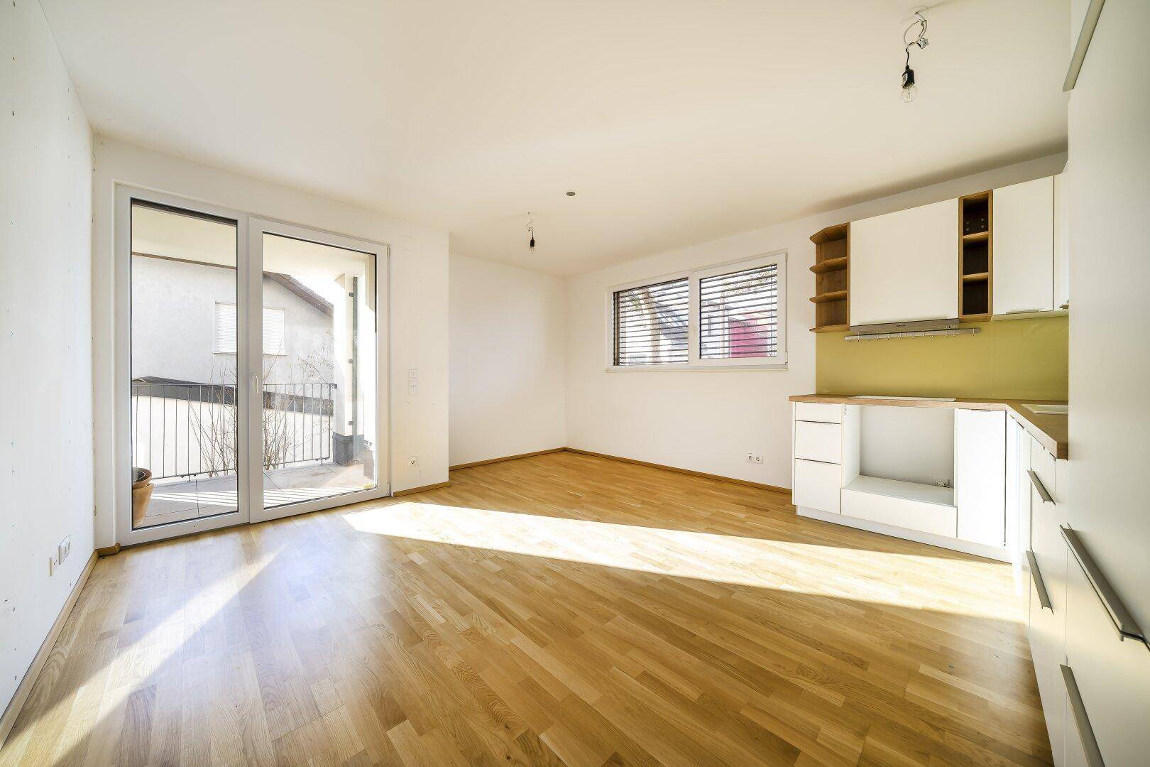 Heller Wohnbereich mit Holzboden und teilweiser Küchenzeile, sonnendurchflutet mit Zugang zum Balkon.