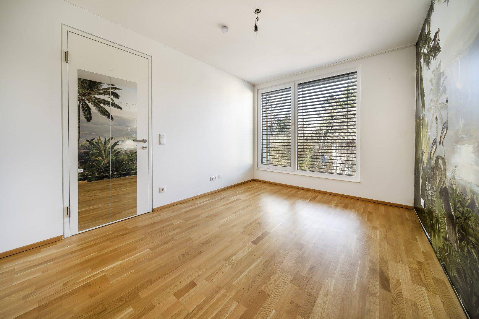 Helles Zimmer mit Holzboden, Fenster mit Jalousien und einer Tür mit Spiegelfläche.