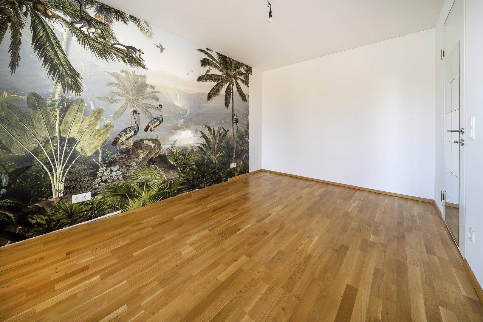 Geräumiges Zimmer mit Holzboden und einer auffälligen tropischen Tapete an einer Wand.